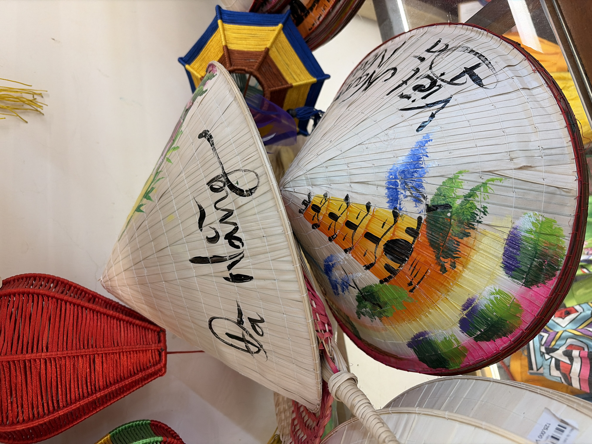 The image displays a close-up view of several conical hats, commonly known as "nón lá," displayed for sale. The hats are made of woven straw and feature decorative elements. One hat in the foreground has a painting of a yellow pagoda surrounded by green foliage with splashes of blue. It is adorned with black calligraphic writing that appears to say "Việt Nam." Another hat above it has black calligraphic writing on its surface, reading "Đà Nẵng." A red lantern made of woven material hangs in the upper left corner, and a colorful, octagonal woven object with yellow and blue threads is visible to the right of the hats. The hats are arranged in a layered manner, suggesting they are part of a market or souvenir shop display. The overall setting appears to be indoors with artificial lighting.