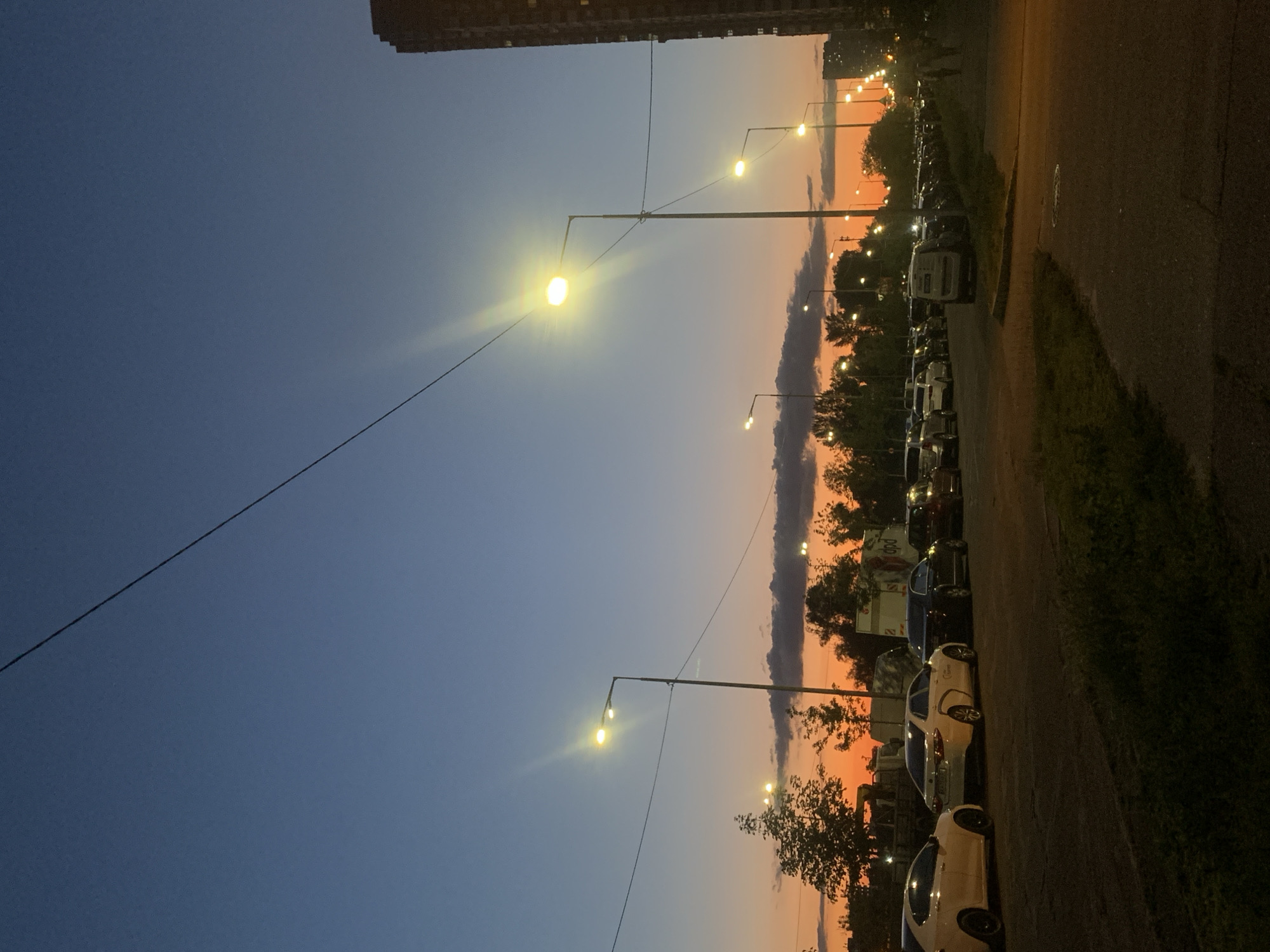 The image depicts a street scene at dusk in Kyiv, Ukraine. The sky transitions from a deep blue at the top to vibrant orange and pink hues near the horizon, indicating the setting sun. Silhouetted against this colorful sky are trees and several streetlights, some of which are illuminated.
In the foreground, a paved road is visible, with a patch of grass and shadows on the left. A line of parked cars stretches along the right side of the road, and some vehicles are also parked on the left. Several buildings, including a tall apartment building on the far right, are visible in the background. The presence of streetlights and buildings suggests an urban environment.
There are no discernible people in the immediate foreground, though a couple of figures can be seen walking on the sidewalk in the far right background. The overall atmosphere is one of evening settling in, with the artificial lights beginning to dominate as natural light fades. The scene appears to be a residential or commercial street with a parking area.