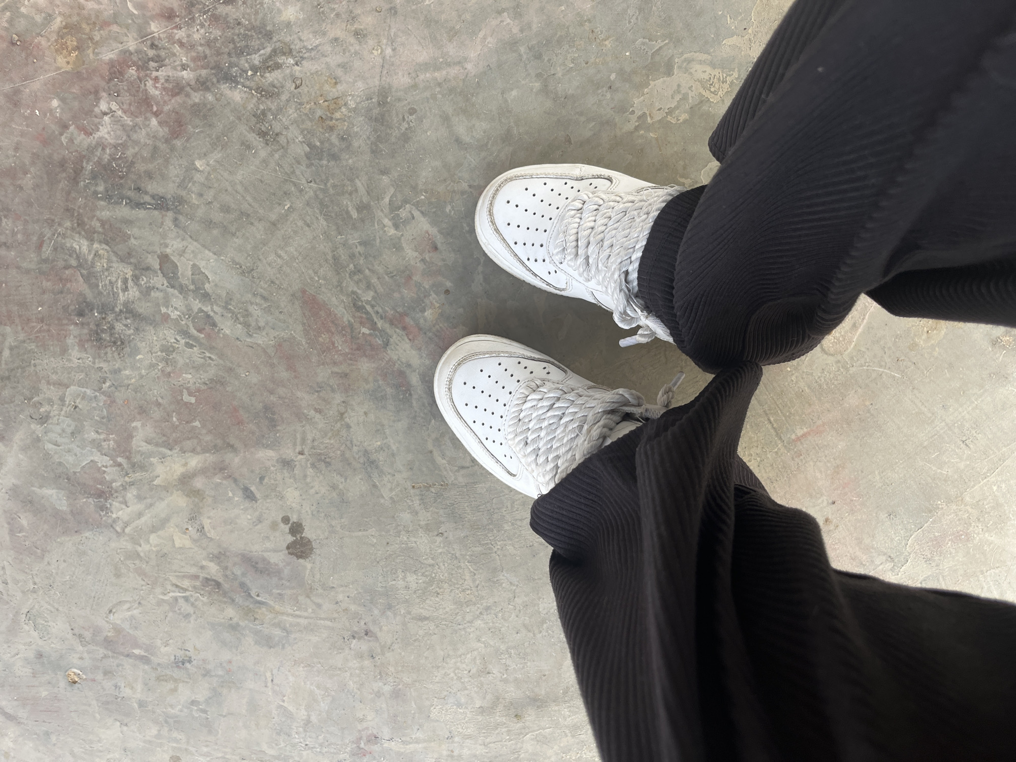 The image depicts a pair of white sneakers with thick rope laces, worn with dark-colored corduroy trousers. The setting is a concrete surface with mottled staining. The location of Juan Griego, Venezuela is not discernible from the image.