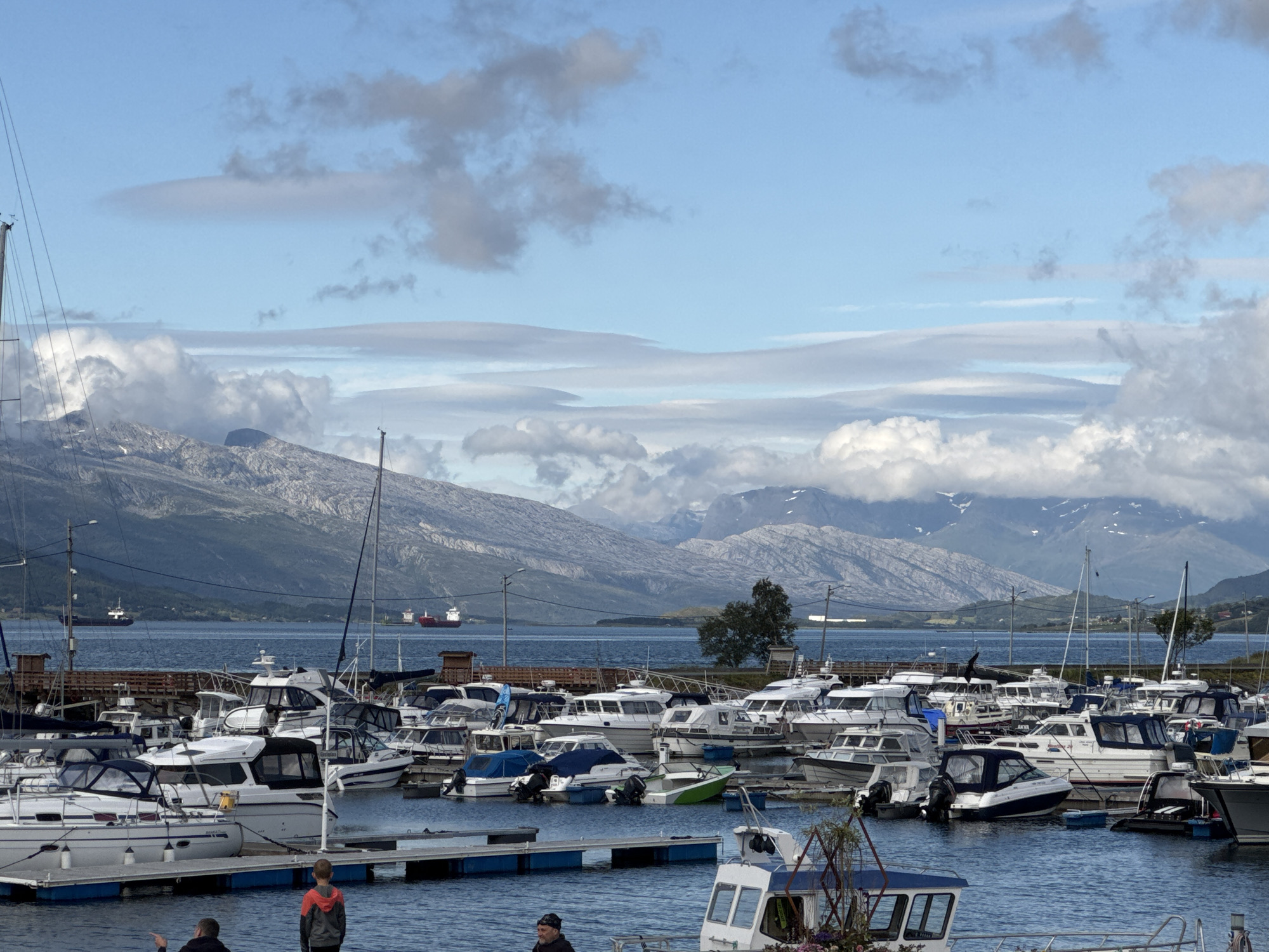 This image depicts a marina filled with numerous boats of varying sizes, docked in a calm body of water. In the foreground, two young boys are standing on a dock, looking out at the water. Behind them, a multitude of motorboats and sailboats are moored. The boats are predominantly white with some featuring blue accents or covers.
Beyond the marina, a large expanse of blue water stretches towards a distant shoreline. In the background, a range of imposing mountains with snow-capped peaks rises dramatically. The sky is a mix of blue with scattered clouds, some of which are dark and appear to be low-lying, suggesting possibly recent or impending weather activity. The lighting suggests it is daytime, possibly late afternoon due to the soft quality of light.
The scene is set in Nesna, Norway, a location known for its coastal beauty and mountainous terrain. The overall atmosphere is peaceful and serene, with the boats resting at anchor and the majestic natural landscape providing a stunning backdrop. There is no visible text in the image.