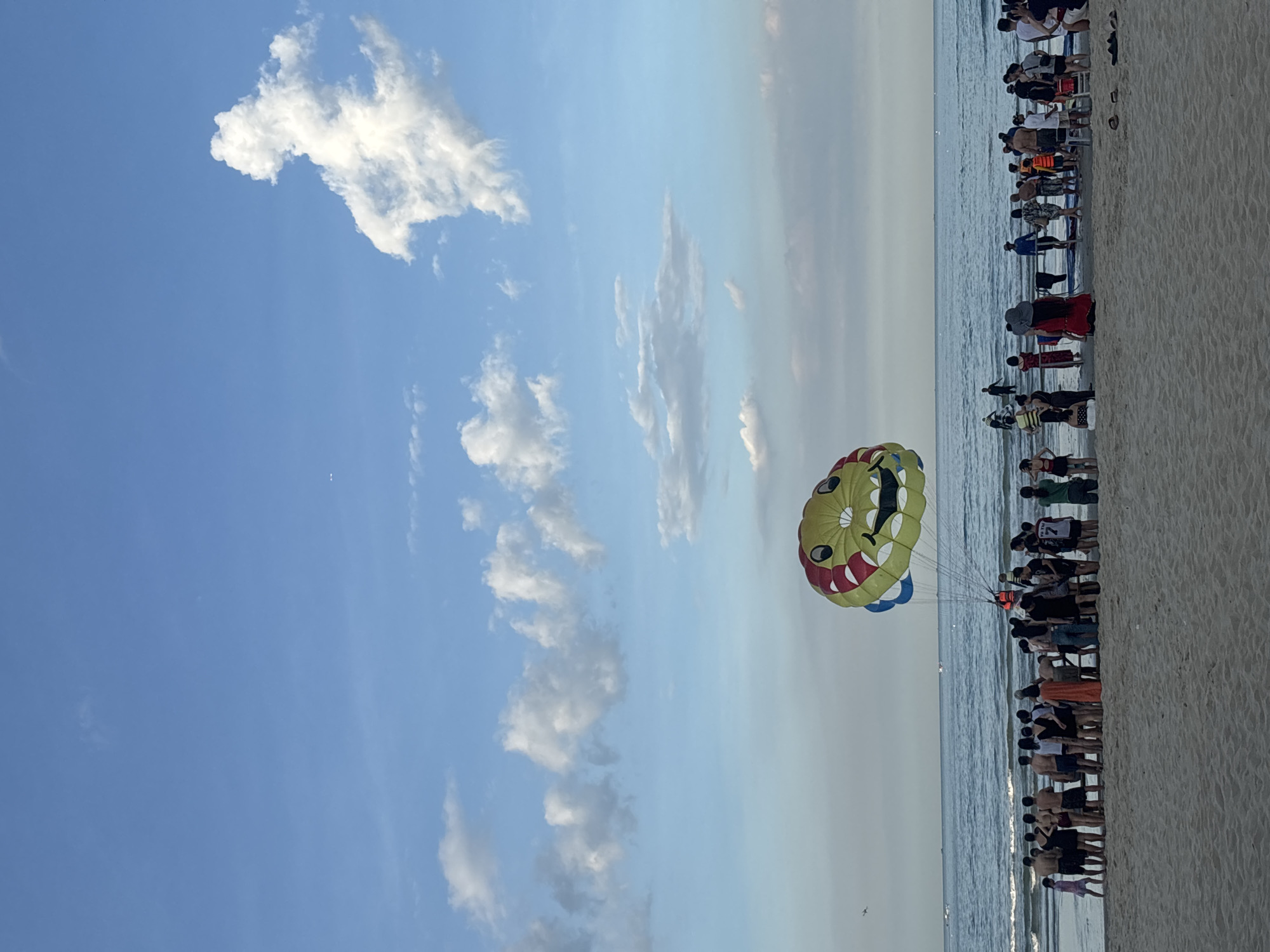This is a photograph of a beach in Đà Nẵng, Vietnam, with a parasail in the shape of a smiling, cartoonish face soaring in the sky above the ocean. The beach is populated with numerous people, many of whom are standing along the shoreline or gathered in groups. The water appears calm with small waves, and the sky is a vibrant blue with scattered white clouds. The parasail is the most prominent object in the air, its colorful design featuring red cheeks and a wide grin. In the distance, several boats are visible on the horizon. The sandy beach is wide and extends towards the water. People are dressed in beach attire, suggesting a warm, recreational atmosphere.