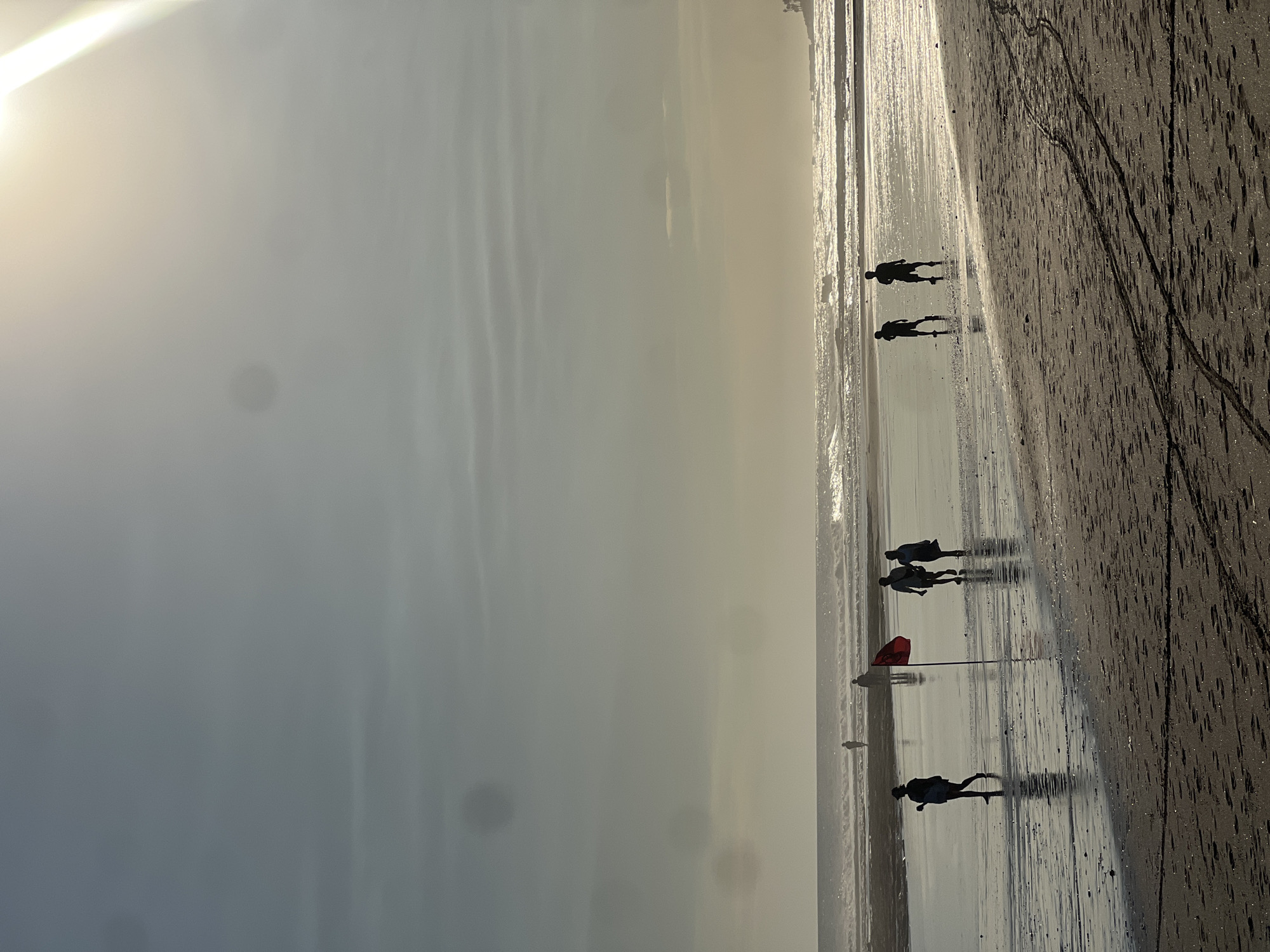 The image depicts a beach scene with several people walking along the wet sand. The sun is low in the sky, casting long shadows, suggesting it is either sunrise or sunset. The sky is hazy, with a bright glare from the sun dominating the upper left portion of the frame. The water's edge is visible, with small waves lapping at the shore. The sand is dark and appears to have been recently wet by the tide, with tire tracks or other markings visible on the surface in the foreground.
There are at least six distinct figures visible, all silhouetted against the bright, reflective wet sand and the hazy sky. Two figures are walking together in the mid-ground, their shadows stretched out behind them. Further back, another group of two or three figures can be seen. Closer to the viewer, a single figure is walking, their shadow also clearly defined. In the distance, a red flag is planted in the sand.
The overall mood is peaceful and serene, with the low light and the natural setting creating a contemplative atmosphere. The location appears to be a beach, likely in a tropical or subtropical region given the brightness of the sun and the clear water. The presence of tire tracks suggests the beach might be accessible by vehicles or that some form of activity has taken place on the sand. There is no visible text in the image.