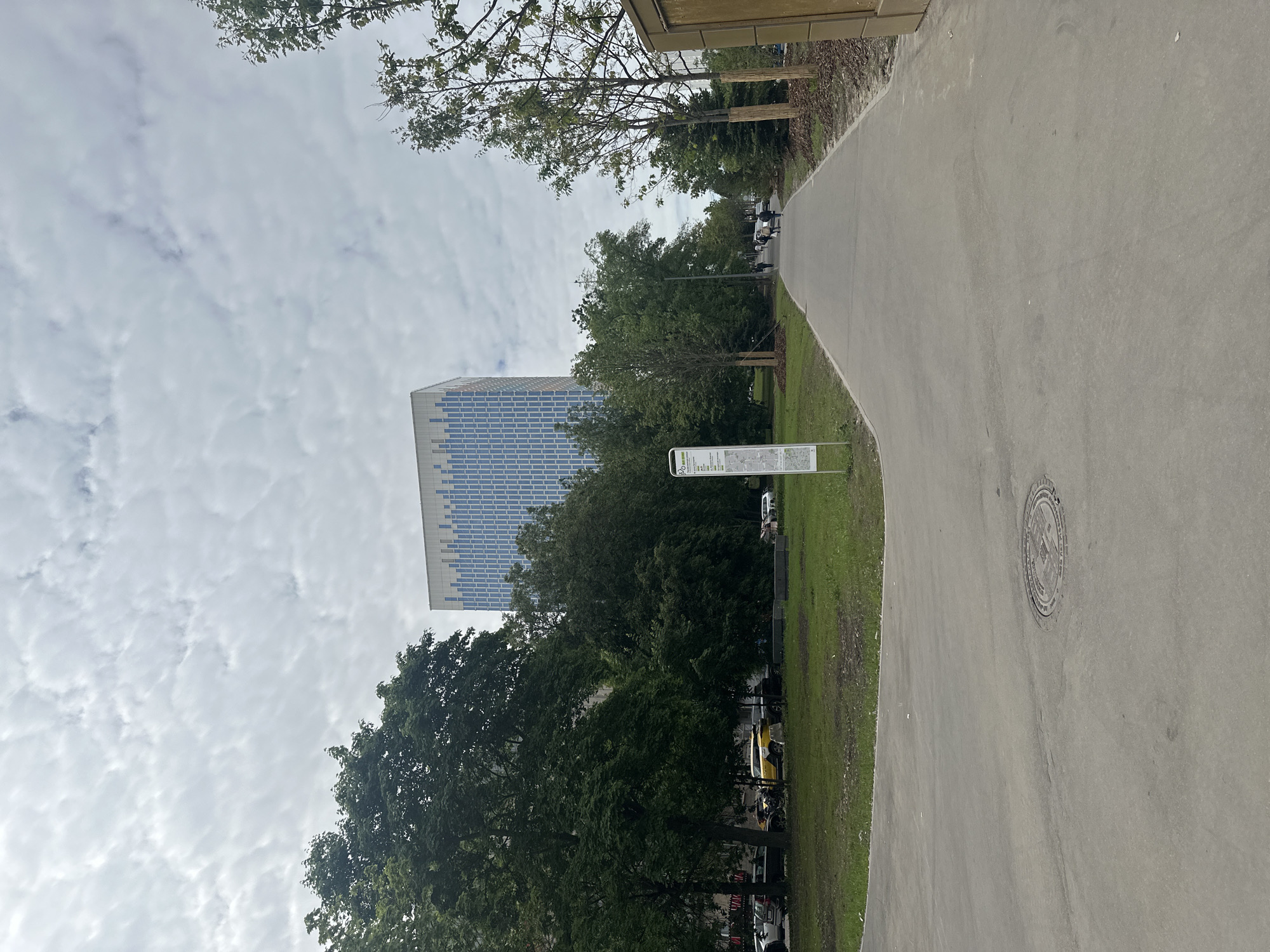 An outdoor scene in Moscow, Russia, captured on an overcast day. The image features a wide, paved pathway that curves from the lower right towards the upper left. On the right side of the path, an informational sign stands upright. The sign is white with green accents, displaying a bicycle icon and text, including "Velo маршруты" (Bike routes) and "ПАРК" (PARK) in Cyrillic, along with map-like graphics. A circular manhole cover is visible on the paved surface in the lower right.
To the left of the path, a stretch of green grass leads to a dense line of mature trees with dark green foliage, which occupy much of the mid-ground. Above the trees and extending into the top corners of the frame, the branches of other trees are visible against a cloudy, light grey sky. In the background, behind the trees and to the left, a tall, modern skyscraper dominates, featuring a distinctive facade composed of a grid of blue, white, and grey rectangular panels or windows. Through gaps in the trees further back, indistinct vehicles, including a yellow one, and what appear to be a few people can be seen, suggesting a parking area or a distant street. The scene depicts a well-maintained urban park or recreational area integrated with modern city architecture.