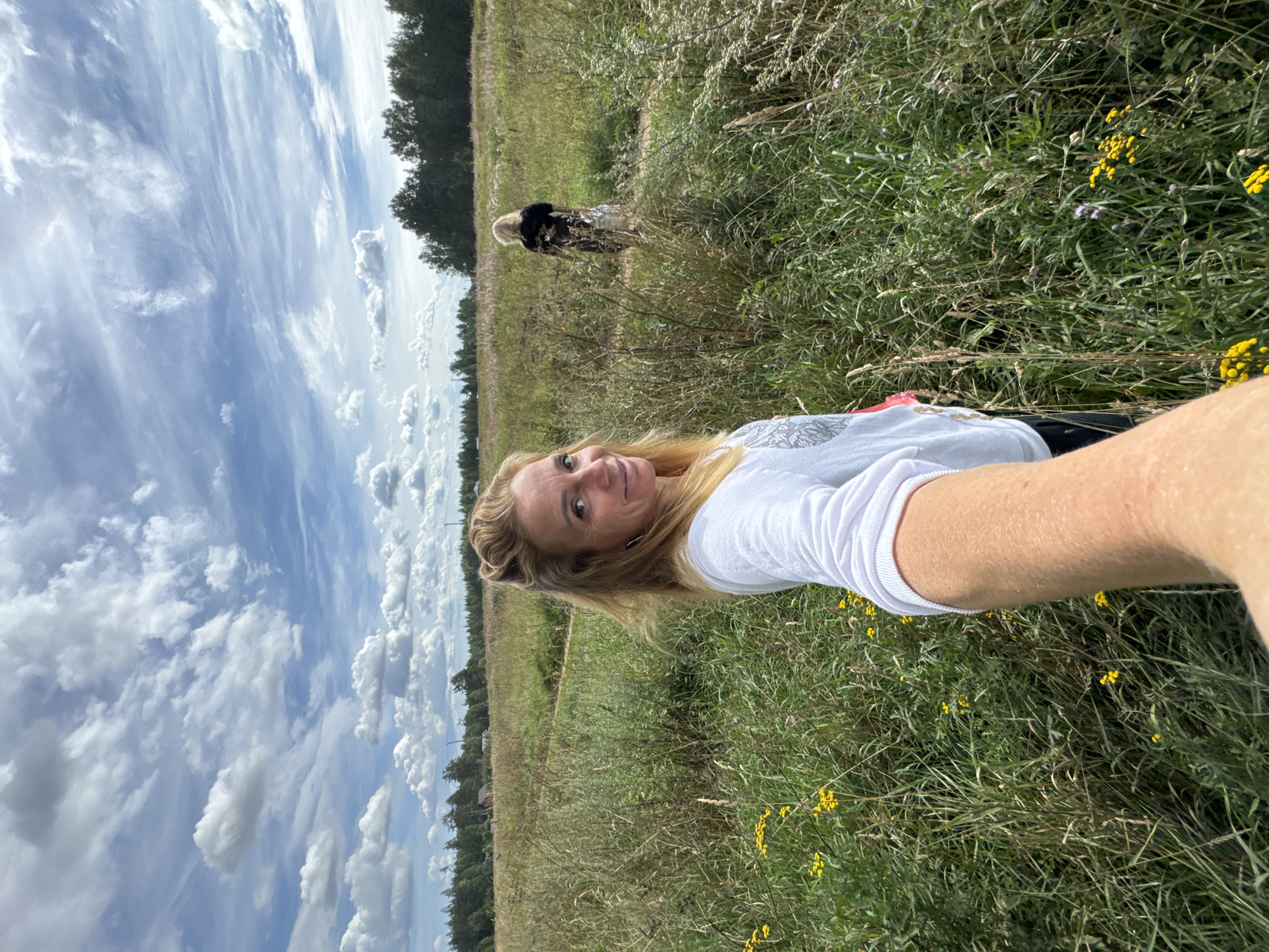 A blonde woman is taking a selfie in a grassy field with a forest in the background. The sky is filled with dramatic, wispy clouds. The woman is wearing a white t-shirt. In the middle ground, a person is standing in the field, facing away from the camera, wearing a black and white outfit. The scene appears to be outdoors, possibly in a rural area of Steklyannyi, Russia, on a day with fair weather. The lighting suggests it is daytime, possibly late afternoon, given the warm tones. The woman's expression is pleasant, as she smiles at the camera. There is no visible text in the image.