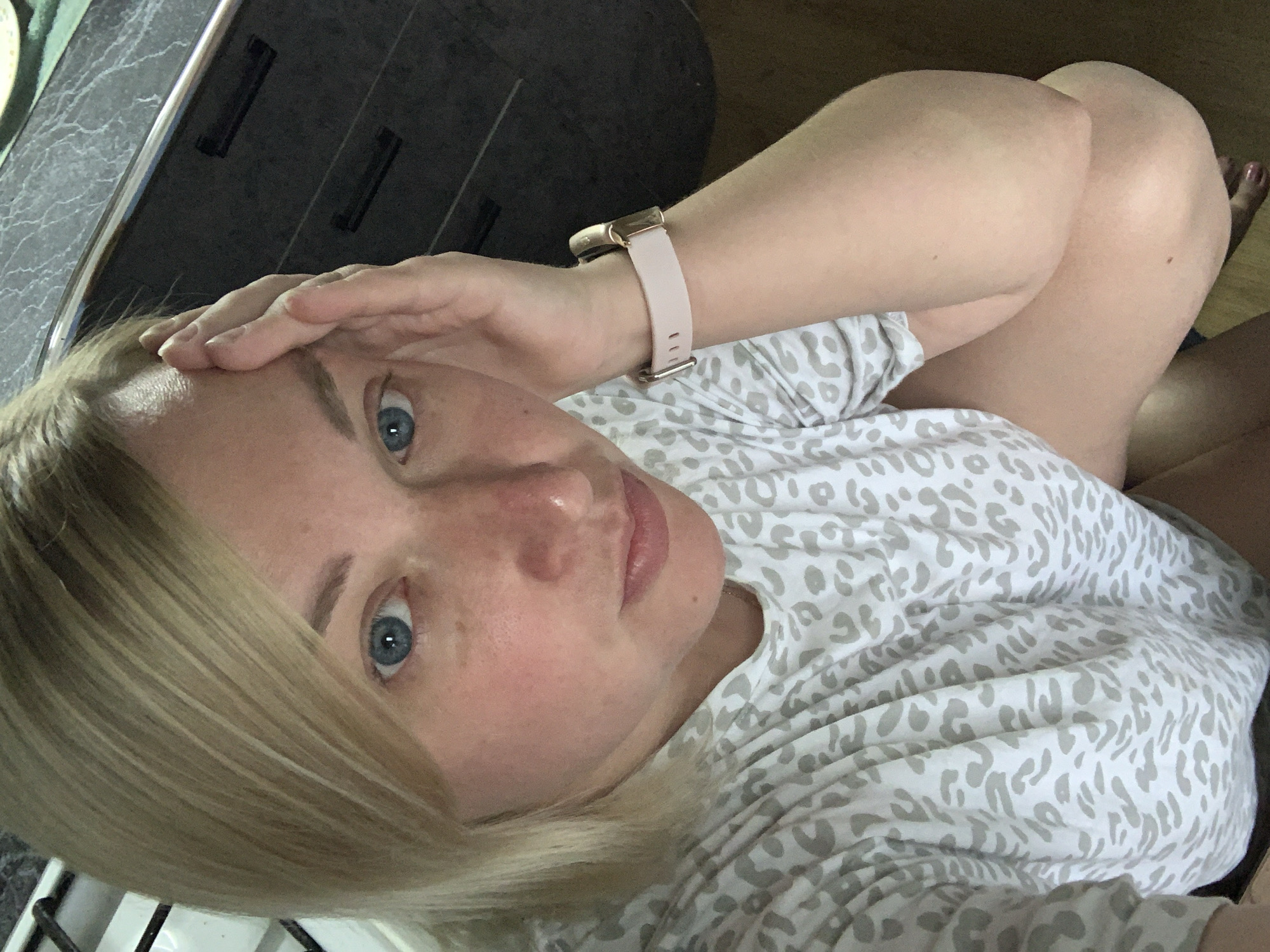 A selfie taken by a blonde-haired woman with blue eyes, who is holding her hand to her forehead. She is wearing a gray and white animal-print shirt and a pink smartwatch. The background includes a dark gray countertop and cabinets, as well as a light brown wooden floor. The location is unknown and there is no evidence of it being the city of Chaikovsky, Russia.