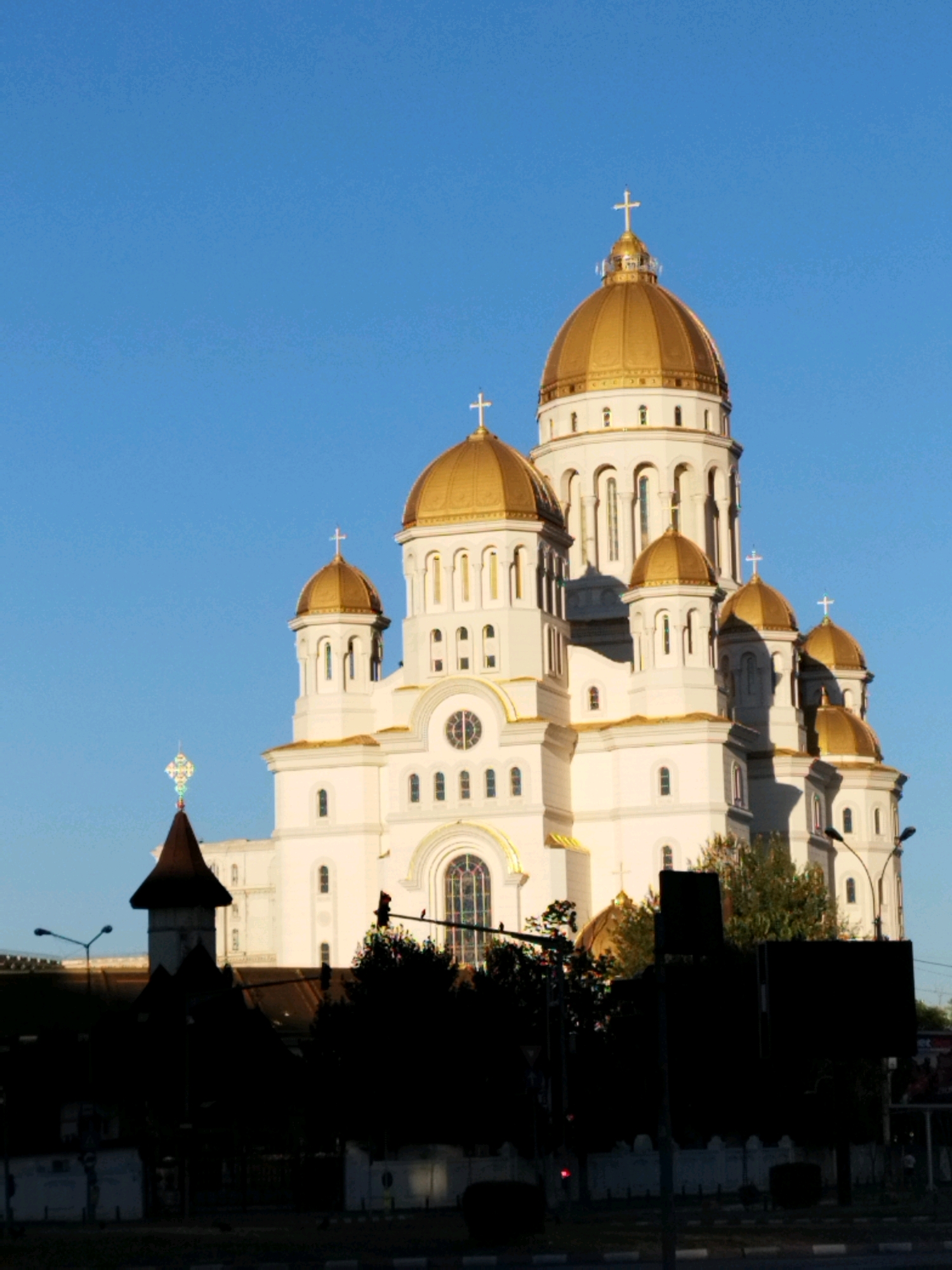 This is an exterior, eye-level, vertical, medium shot of the Patriarchal Cathedral of Saints Constantine and Helena in Bucharest, Romania, taken in daylight. The massive cathedral, built of white stone, features multiple golden domes, several of which are topped with crosses. The largest dome is centrally located and dominates the upper right portion of the frame. The building exhibits classic Orthodox architectural elements, with arched windows and decorative cornices. In the foreground, silhouetted trees and the dark shapes of urban structures, including streetlights and what appear to be billboards or bus stops, obscure the lower part of the scene. The sky is a clear, vibrant blue. The lighting suggests it is either early morning or late afternoon, with the sun casting long shadows. No people are visible in the image.