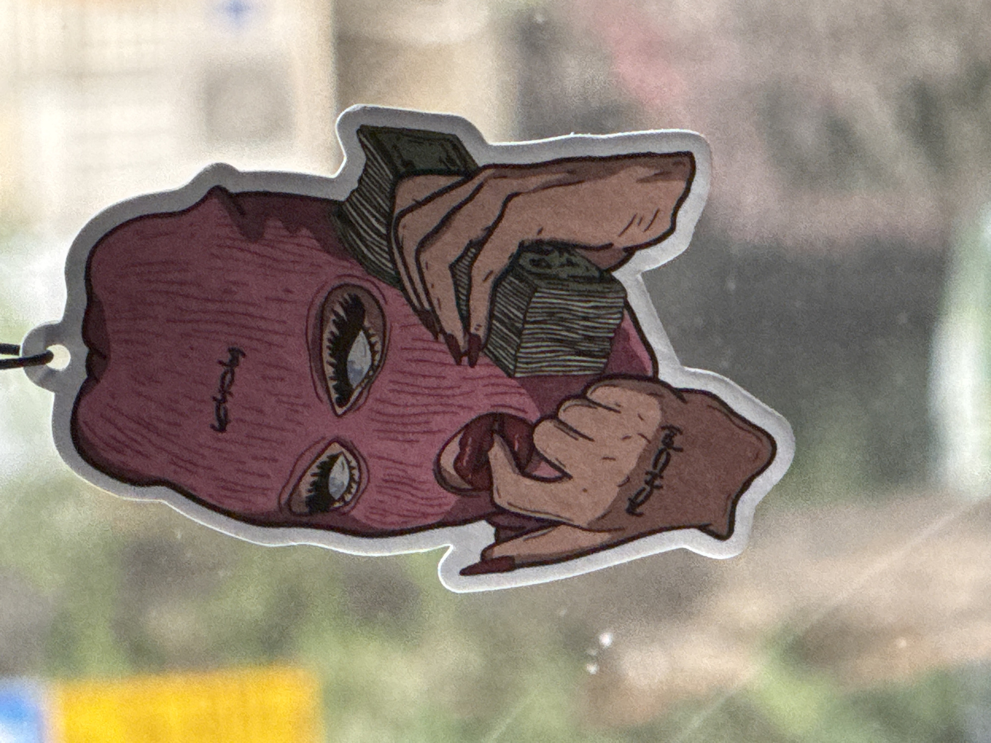 The image shows an air freshener sticker featuring a stylized illustration. The illustration depicts a person wearing a pink ski mask, with visible eyes. The person is holding stacks of money in both hands. The sticker is white-bordered and has a small hanging loop at the top. The background appears to be an out-of-focus view through a window, possibly a street scene. The image does not provide any direct information to identify the location as Ramat Gan, Israel.