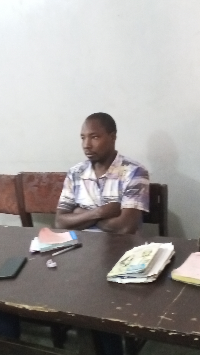 The scene, located in Dakata, Nigeria, depicts an adult male seated at a dark brown table. He has short dark hair and visible facial stubble, wearing a short-sleeved, multi-patterned collared shirt. His arms are crossed over his chest, and his gaze is directed towards the left side of the frame. Behind him, two dark brown wooden chairs are partially visible against a plain, light-colored wall. On the table, from left to right, are a dark smartphone, a small stack of papers or documents with a pen resting on them, and a larger stack of currency notes. No direct interaction is occurring within the frame.