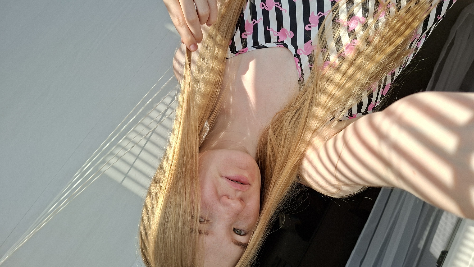 A young woman with long, blonde hair and freckles is lying down and taking a selfie. She is wearing a black and white striped top with pink flamingos on it. The sunlight is casting shadows across her face and body, creating a striped pattern. The background appears to be indoors, with white blinds or curtains visible. There are no discernible Chaikovsky, Russia landmarks or context in this image.