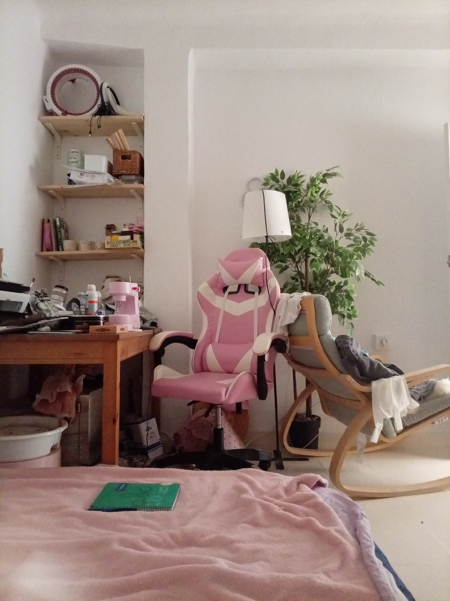 The image depicts an indoor scene, possibly a bedroom or living area, characterized by a collection of objects suggesting a lived-in space. A prominent pink and white gaming chair is positioned in the center-right of the frame. To its left, a wooden desk is cluttered with various items, including what appears to be a small pink sewing machine, bottles, and other miscellaneous objects. Above the desk, a shelf unit displays more items, such as books, containers, and a basket.
In the background, to the right of the gaming chair, a rocking chair with clothing draped over it is visible, and behind it, a large artificial plant stands beside a floor lamp with a white shade. In the foreground, a light pink blanket or throw covers a surface, with a green notebook resting on it. The walls are painted white, and the flooring appears to be light-colored tile or linoleum.
This scene does not provide any specific visual cues that would allow for its localization in Olula del Río, Spain, or any other city. The interior setting is generic, lacking architectural or decorative elements that are distinctly Spanish or specific to that particular municipality.