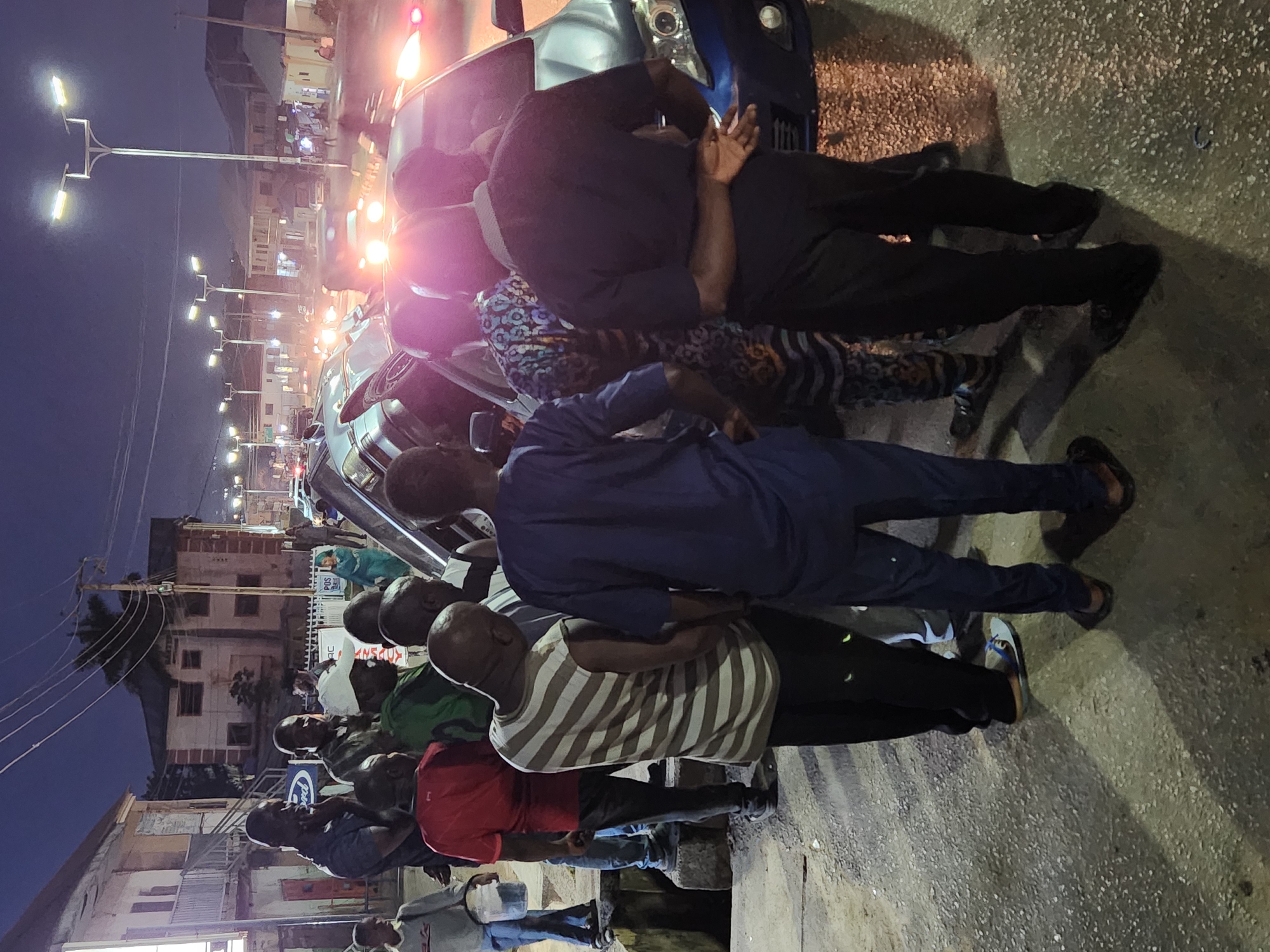 A group of people are standing and some are laying on the ground next to a car at night. The location appears to be urban with streetlights and buildings in the background. The city is unknown.