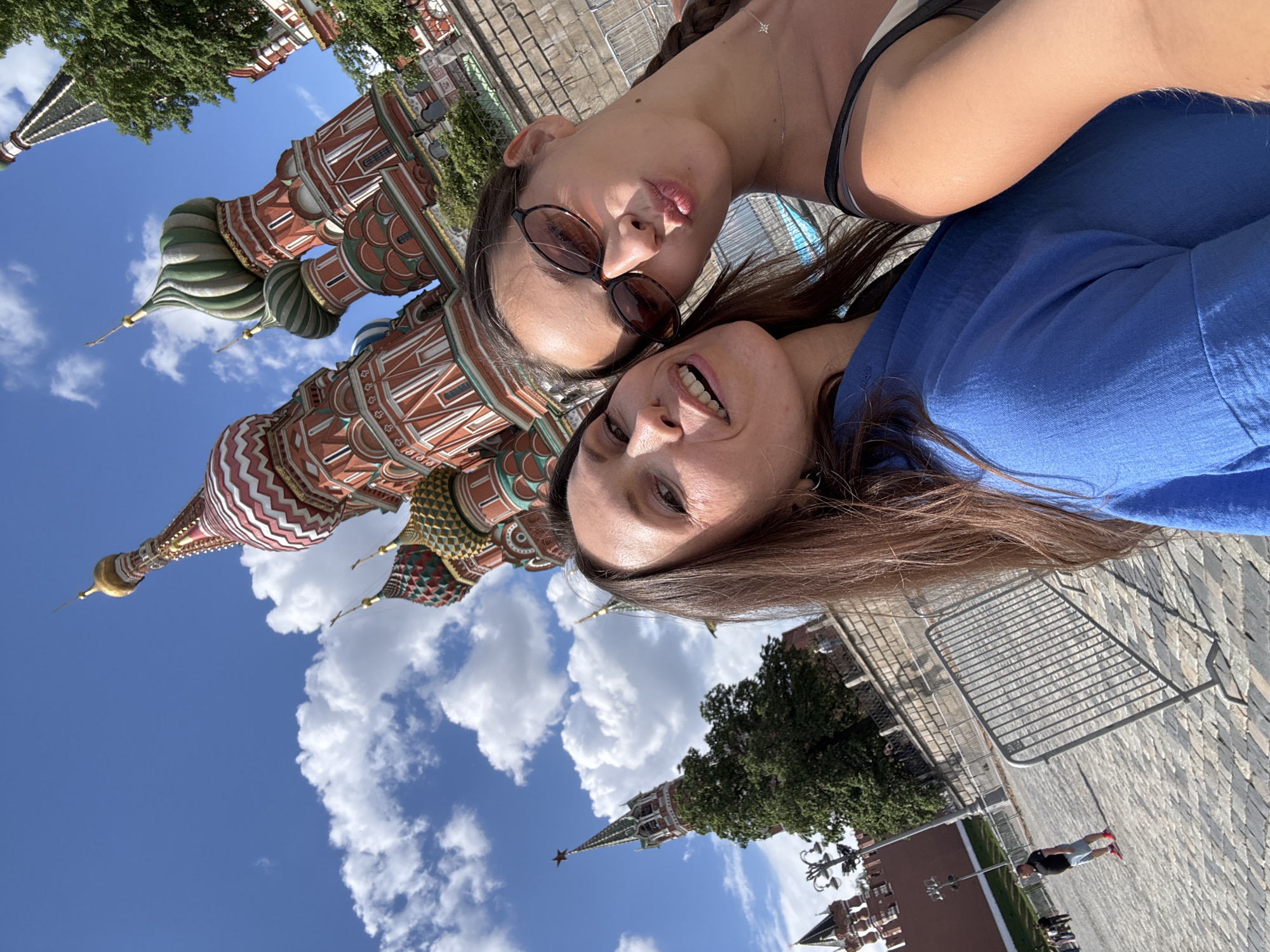 Two women pose for a selfie in Moscow, Russia. Behind them is St. Basil's Cathedral with its distinctive onion domes and the Kremlin with its red star-topped spire. The sky is blue with scattered clouds. A metal barricade is visible in the background, along with trees and a pedestrian.