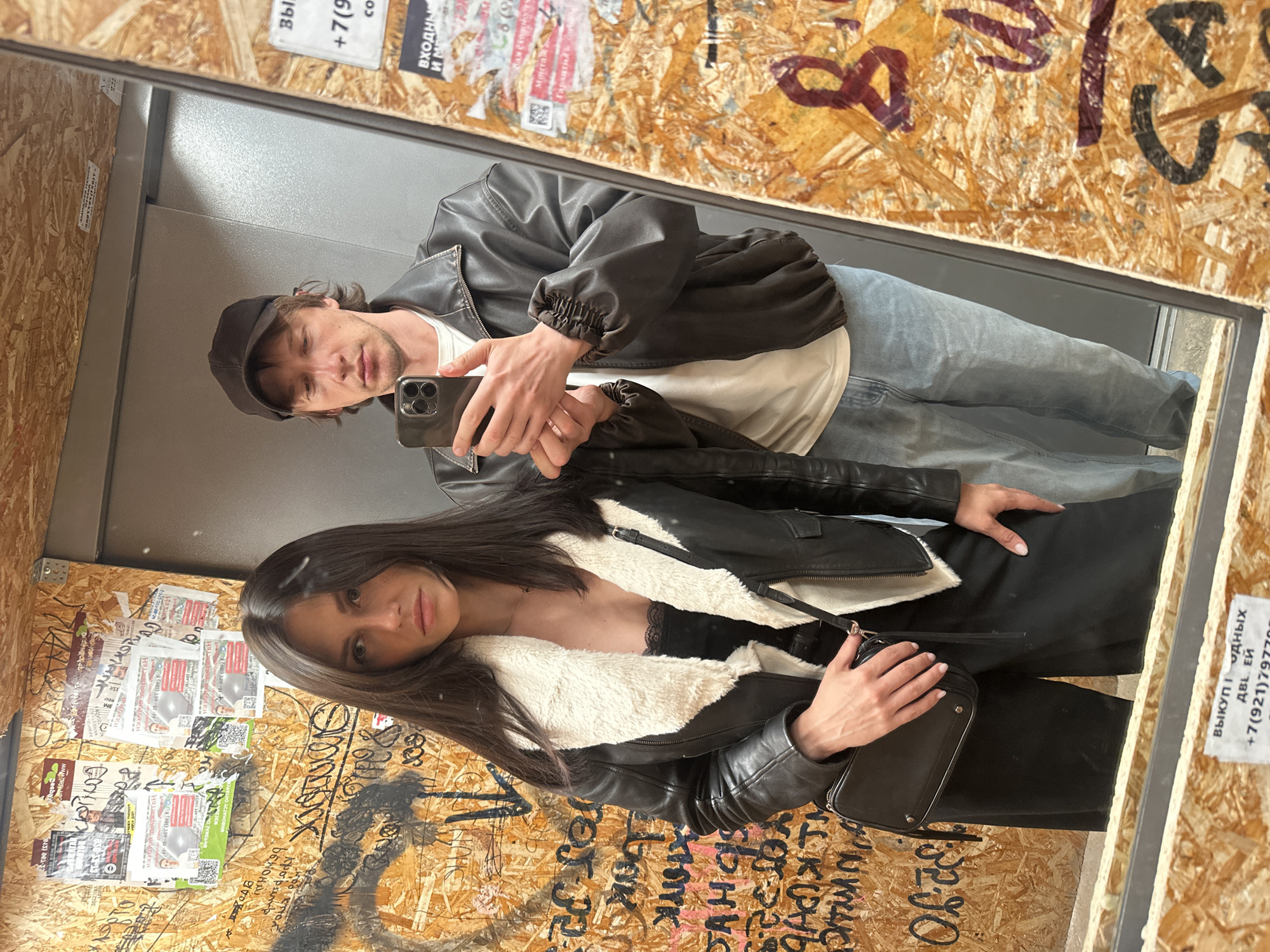 A young couple is taking a mirror selfie in what appears to be an elevator. The woman, on the left, has long dark hair and is wearing a black leather jacket with a white fur collar, a black lace top, and black wide-leg pants. She is carrying a black crossbody bag. The man, on the right, is wearing a dark baseball cap, a dark leather jacket, a white t-shirt, and light-wash jeans. He is holding a smartphone to take the picture. The elevator walls are covered in graffiti and posters, giving the scene a gritty, urban feel. The lighting is harsh and direct, characteristic of indoor artificial lighting.