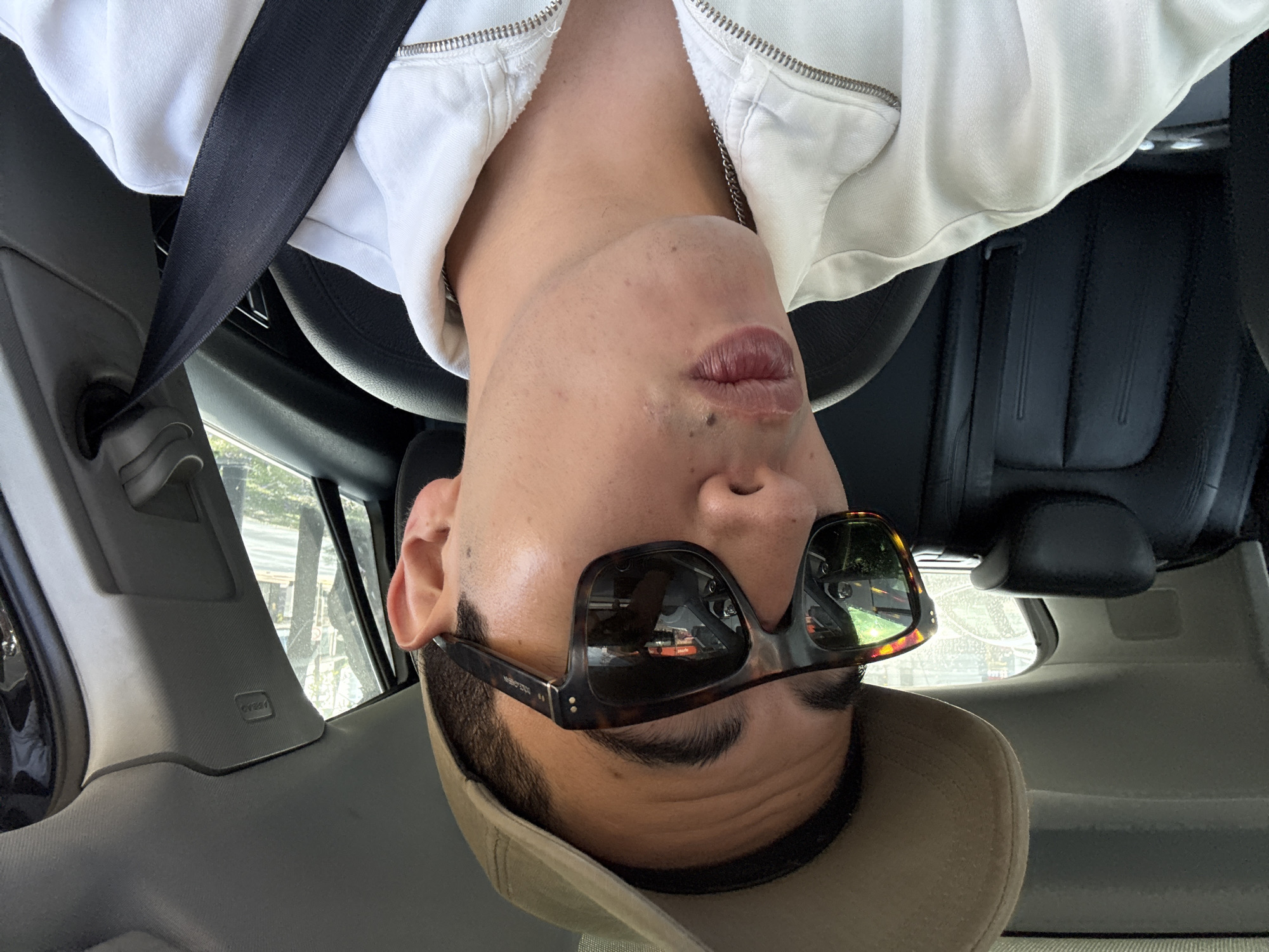 A close-up, portrait-oriented shot depicts a person seated in the front of a car during the daytime in Puebla, Mexico. The image is taken from a low angle, looking upwards at the subject. The person, who appears to be male, is wearing a white hooded jacket with a prominent silver zipper, a black seatbelt across their chest, and a beige or light brown baseball cap. They are also sporting tortoise-shell framed sunglasses that reflect elements of the car's interior and what lies ahead, including a hand or arm possibly belonging to the photographer or the person themselves. The sunglasses have the text "VINTAGE EYEWEAR" visible on the temple. The subject's lips are slightly pouted, suggesting a deliberate pose, and they have some facial stubble and small moles on their chin and cheek. A silver chain is visible around their neck.
The car's interior features dark-colored, likely black, leather seats and a gray or dark-toned dashboard and door panels. A black steering wheel is partially visible on the left. Through the car windows, blurred foliage and what appear to be urban structures can be seen in the background. A prominent "AIRBAG" label is visible on the car's interior pillar near the side window. The overall lighting is bright, indicative of daylight.