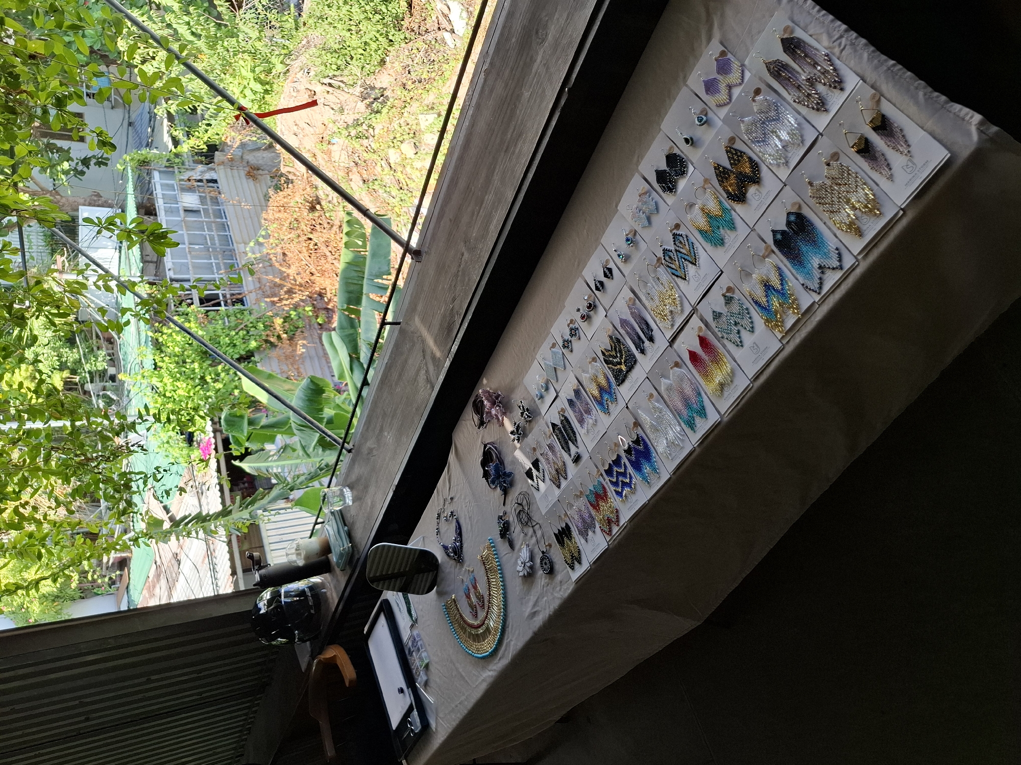 The image depicts a tabletop display of handmade beaded earrings and necklaces, set up on a light-colored tablecloth. The table is positioned against a window overlooking an outdoor scene with lush green foliage, residential buildings with green roofs, and some dry brush. The window has a wooden frame and a metal railing.
On the table, the jewelry is arranged on white card stock backing, creating rows of colorful earrings. There are various styles and colors, including chevron patterns, teardrop shapes, and fringe designs. Some necklaces are also visible, including a prominent gold and turquoise beaded necklace on the left side of the table, and a darker beaded necklace to its right. Other items on the table include a small mirror, a black container with small compartments holding what appear to be beads or jewelry findings, and a pair of scissors. On the windowsill above the table, there are a black thermos, a glass with ice and liquid, and a small glass of water.
The outdoor view from the window suggests a tropical or subtropical environment, likely in a city like Nha Trang, Vietnam, given the presence of dense vegetation and residential structures common in such locations. The sunlight filtering through the leaves indicates it is daytime. The overall impression is that of an outdoor market stall or a display set up in a cafe or shop with an open-air view. No people are visible in the image.