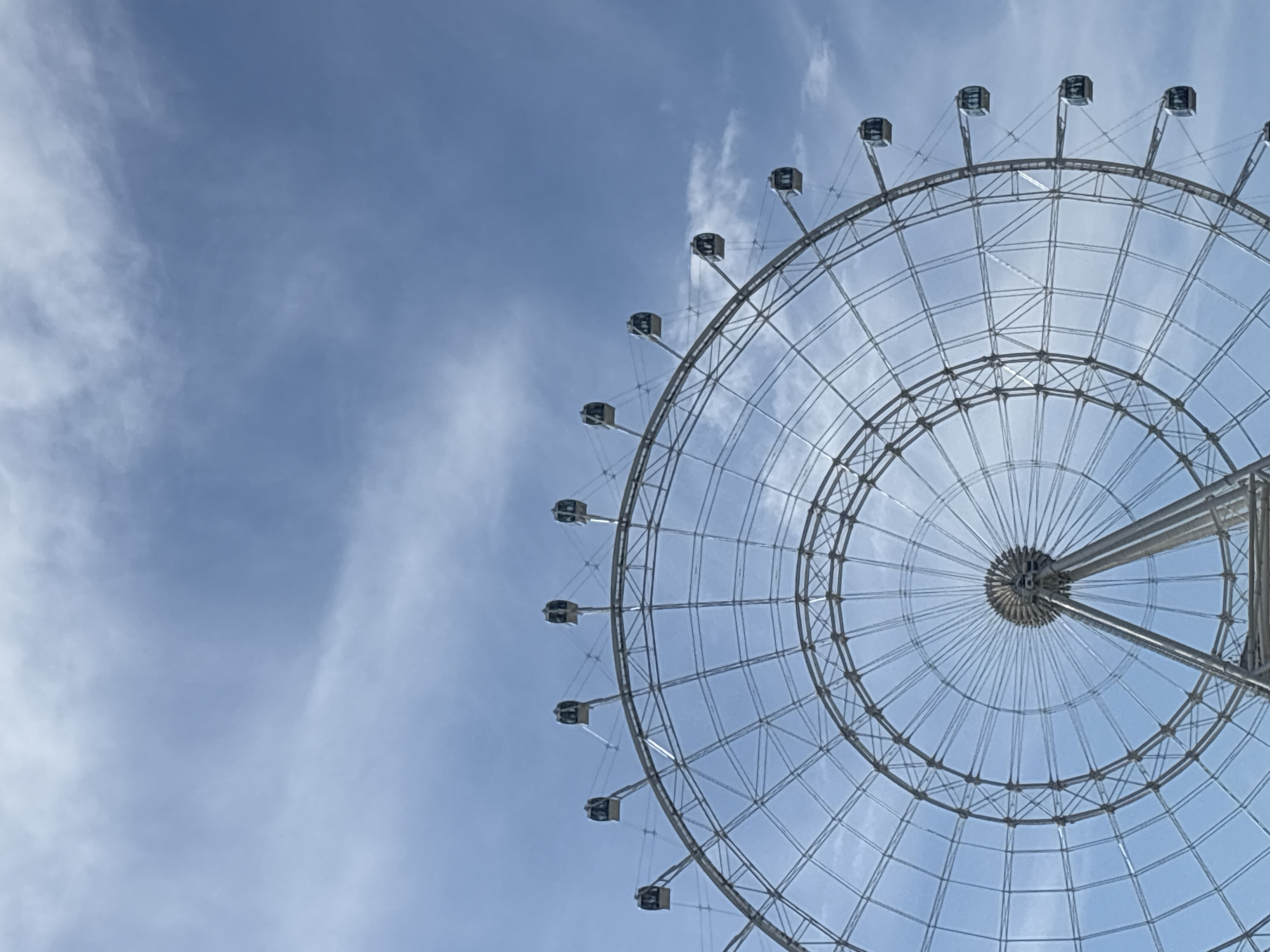 A low-angle, full shot captures a towering Ferris wheel against a pale blue sky with wispy clouds. The Ferris wheel, constructed of silver metal, dominates the lower half of the frame. Its intricate lattice structure is clearly visible, with numerous spokes radiating from the central hub and connecting to the outer rim. Suspended from the rim are several enclosed, rectangular cabins, each with large windows. The cabins are spaced evenly around the wheel, with a few visible at the top and along the right side. The sky above is a soft blue, with delicate white clouds scattered across it, creating a sense of open space and light. The image is sharp and clear, focusing on the architectural details of the Ferris wheel and the expansive sky.