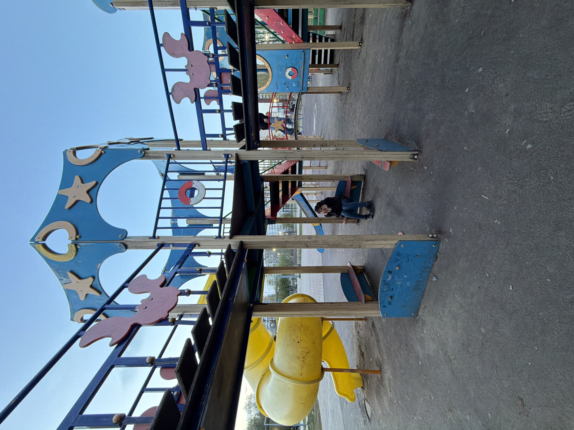 The image shows a playground in Chaikovsky, Russia. The playground has a complex structure with slides, ladders, and decorative elements like starfish and seahorse cutouts. A person in dark clothing and jeans is sitting or crouching near the playground equipment. The ground is paved, and the sky is visible in the background.