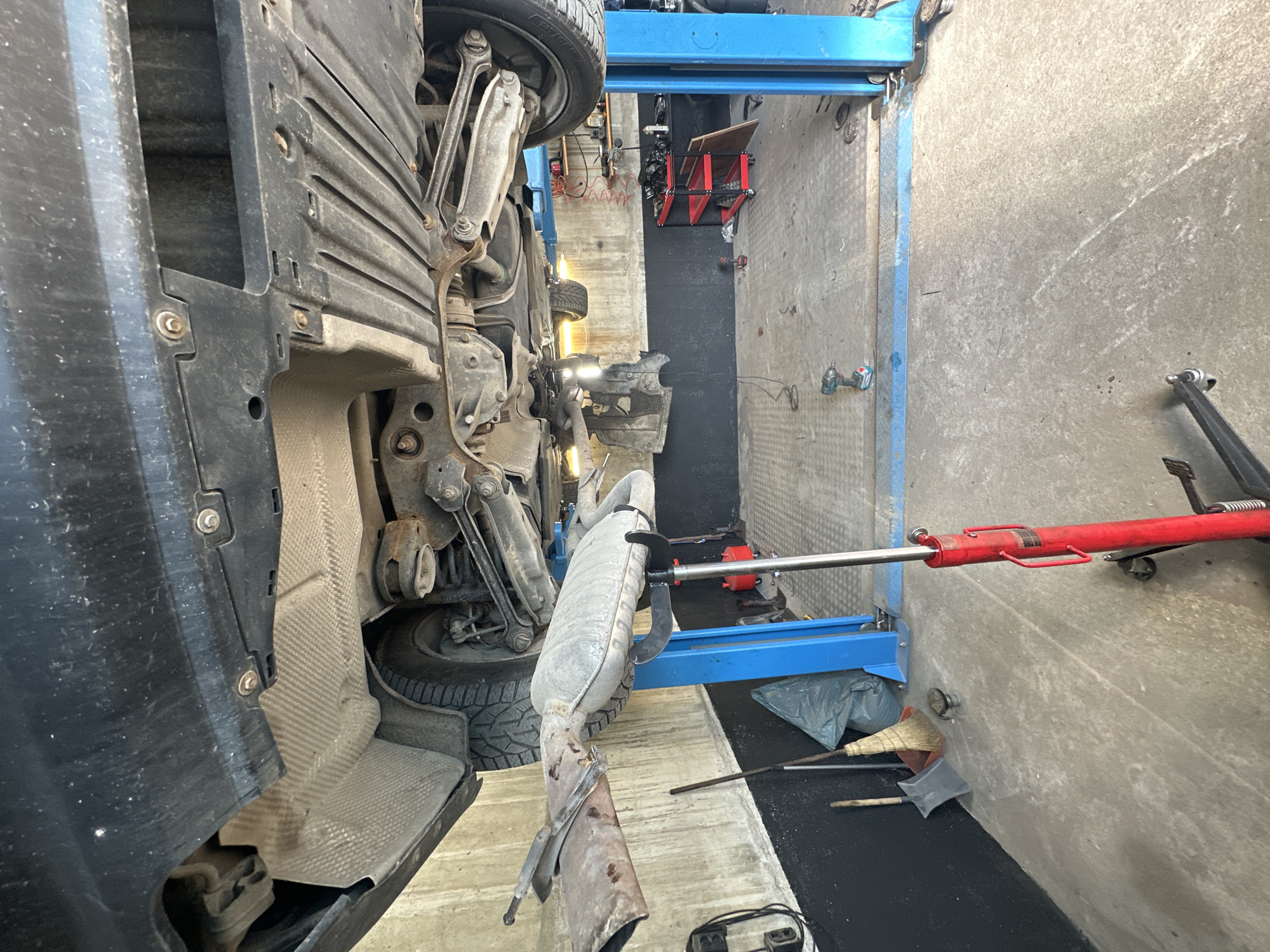 The image depicts the underside of a car being serviced in an auto repair shop. A blue hydraulic lift is holding the vehicle, revealing its exhaust system, suspension, and undercarriage. A red floor jack is also visible, positioned beneath the exhaust. The shop floor appears to be concrete, with various tools and equipment scattered around, including a broom, a trash bag, a red cart with parts, and power tools. The walls are concrete and painted black in the lower section. The scene suggests ongoing maintenance or repair work on the vehicle. The location is identified as an auto repair shop in Kazanlak, Bulgaria. There are no people visible in the image.