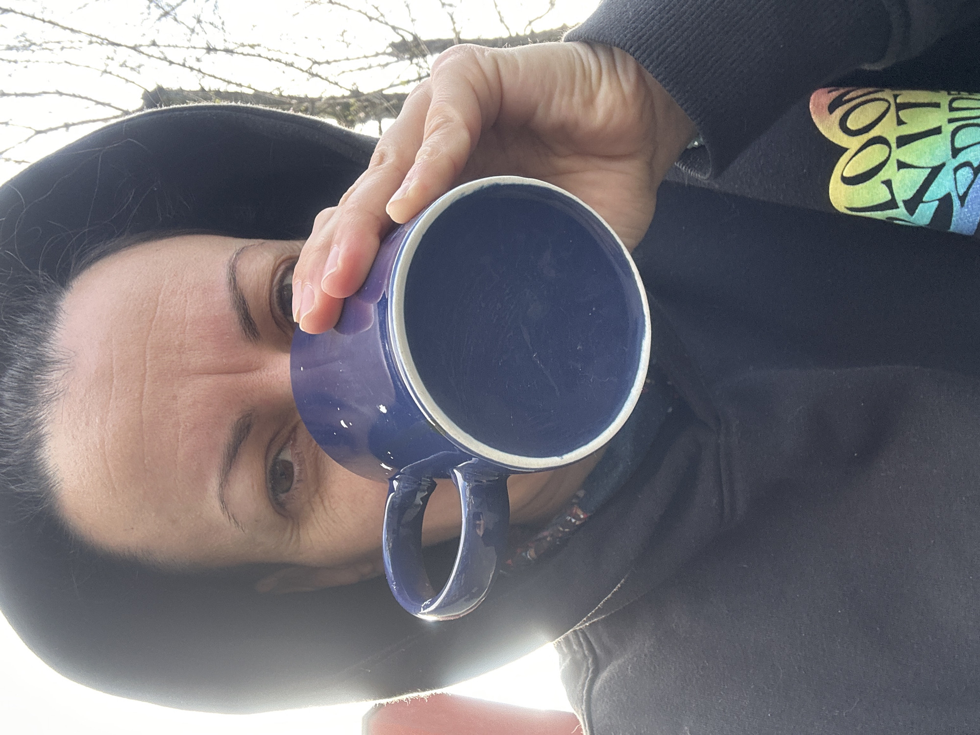 A person is holding a blue mug with a white rim, close to their face. The person is wearing a black hooded sweatshirt. The inside of the mug is dark blue. The person's hand is holding the mug. The background shows blurry branches and bright light, suggesting an outdoor setting. The sweatshirt has colorful text on the sleeve. There is not enough information to place the image in Sofia, Bulgaria.