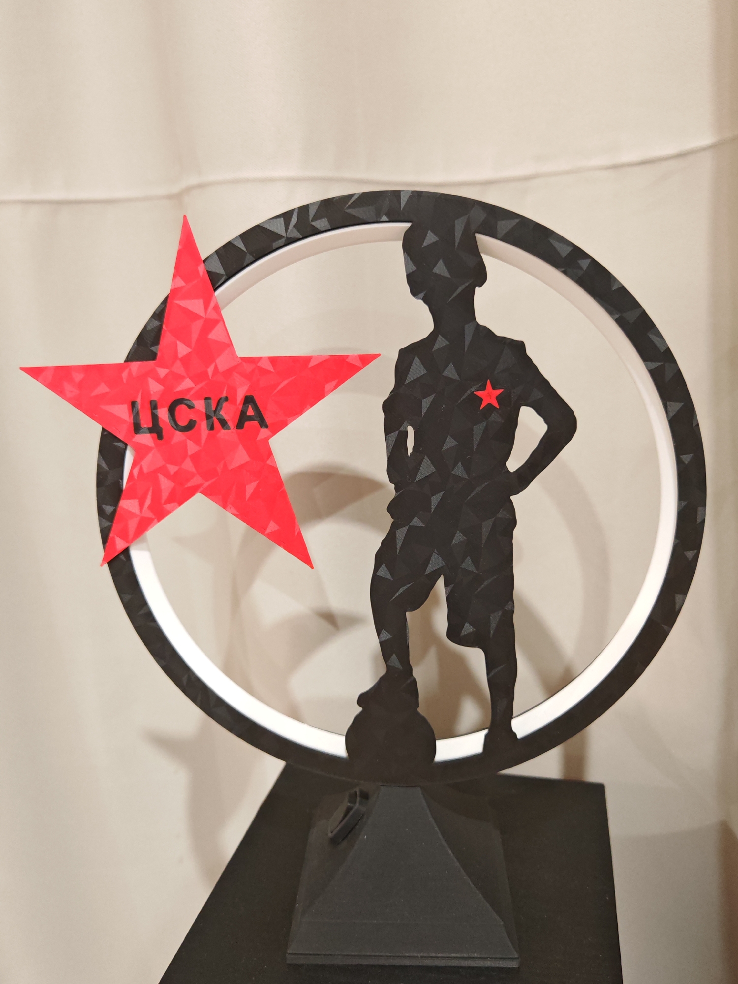 The image shows a trophy with a black silhouette of a standing person inside a circular frame. The person is depicted with one hand on their hip and is wearing shorts. A small red star is placed on the person's chest. To the left of the silhouette, a large red five-pointed star is attached, with the Cyrillic letters "ЦСКА" inscribed in black on its surface. The trophy is mounted on a black pyramidal base. The background consists of a draped, light-colored fabric, possibly a curtain or wall hanging. The lighting appears to be diffused, casting soft shadows. There is no identifiable location in the city of Plovdiv, Bulgaria, depicted in this image.