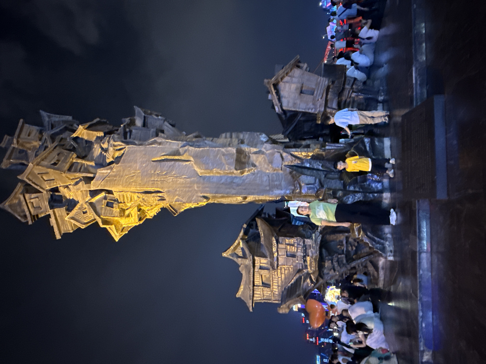 At night, a group of people are gathered around a large, abstract sculpture that resembles a cluster of stacked houses. The sculpture is illuminated by warm lights, casting a golden glow on its textured surfaces. In the foreground, a woman in a green t-shirt and black pants stands to the right of the sculpture. To the left of her, a young boy in a yellow shirt and black pants stands with his hands by his sides. In the background, several other people are visible, some sitting and others standing, engaged in conversations or taking pictures. The scene is set against a dark sky, with city lights visible in the distance. The sculpture is located in Yuzhong District, China.