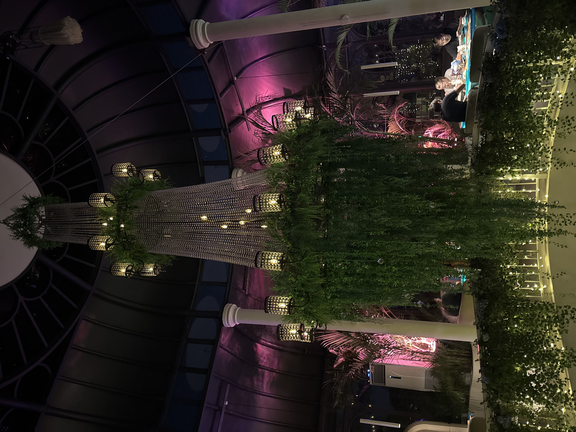 The image depicts an indoor dining area with a large, elaborate chandelier adorned with greenery and multiple lights. The chandelier hangs prominently in the center of the frame, casting a warm glow. In the background, several people are seated at tables, engaged in dining. The environment appears to be a restaurant or a similar establishment, characterized by ambient lighting, plants, and architectural features like columns and a domed ceiling. The overall atmosphere is dimly lit and sophisticated. The location is identified as North Kuta, Indonesia.