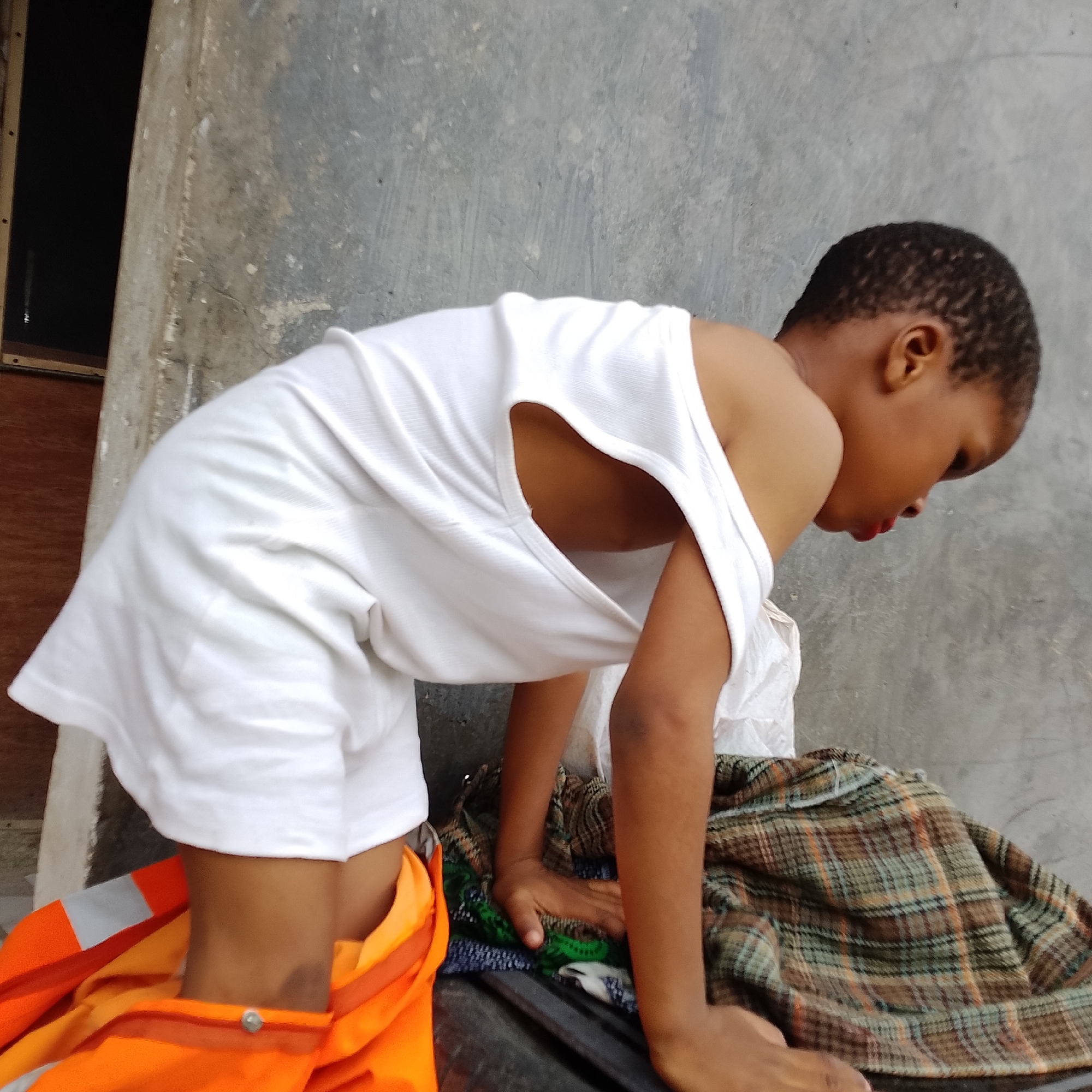 A child is bent over a collection of fabric, with a concrete wall in the background. The child is wearing a white tank top and orange reflective clothing around their waist. One hand is resting on the fabric, while the other is not visible. The fabric appears to be a plaid blanket or textile. The scene suggests an outdoor environment, possibly in a residential or industrial area within Ado, Nigeria. The lighting is natural, indicating daytime.