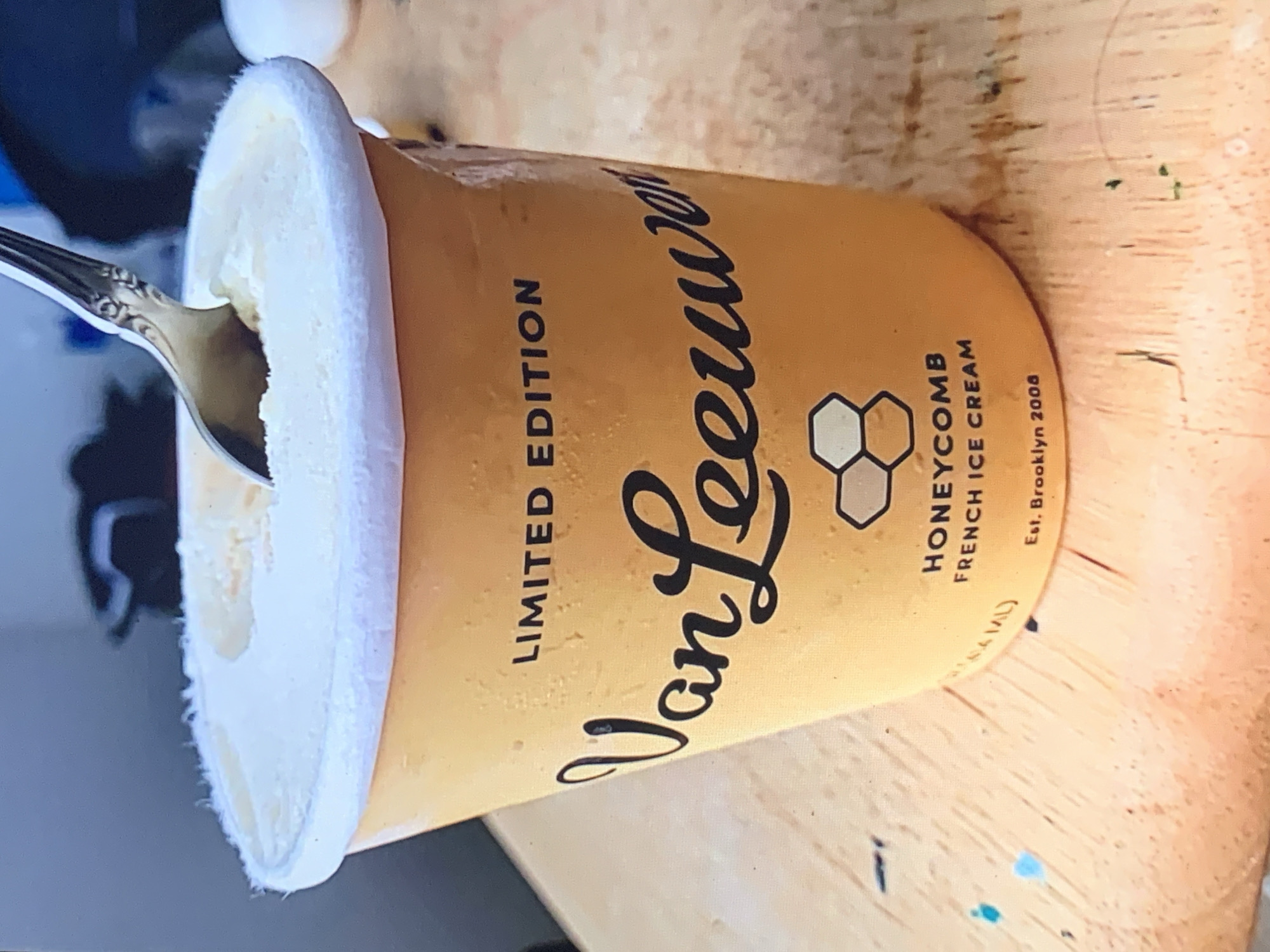 The image displays a pint of "Van Leeuwen" ice cream, oriented horizontally on a light-colored wooden surface. A silver spoon with an ornate handle is partially inserted into the creamy, pale yellow ice cream, suggesting it is being eaten or is about to be consumed.
The ice cream pint is primarily light yellow or beige. Prominently featured on its label is the brand name "Van Leeuwen" in a stylish, black script font. Above this, the text "LIMITED EDITION" is printed in sans-serif capital letters. To the right of the brand name, a logo depicting interconnected hexagons, symbolizing a honeycomb, is visible. Below the logo, the label specifies the flavor as "HONEYCOMB FRENCH ICE CREAM," followed by "Est. Brooklyn 2008." A small, partially visible text likely indicates the volume, appearing as "(ML)". The ice cream fills the pint, with some slight melting visible where the spoon has entered.
The background is a textured wooden surface, possibly a table, which appears to have a few small, faint green and blue specks on it. The overall scene suggests a casual indoor setting, likely at home. No people are visible, and no specific time of day or emotional cues are discernible.