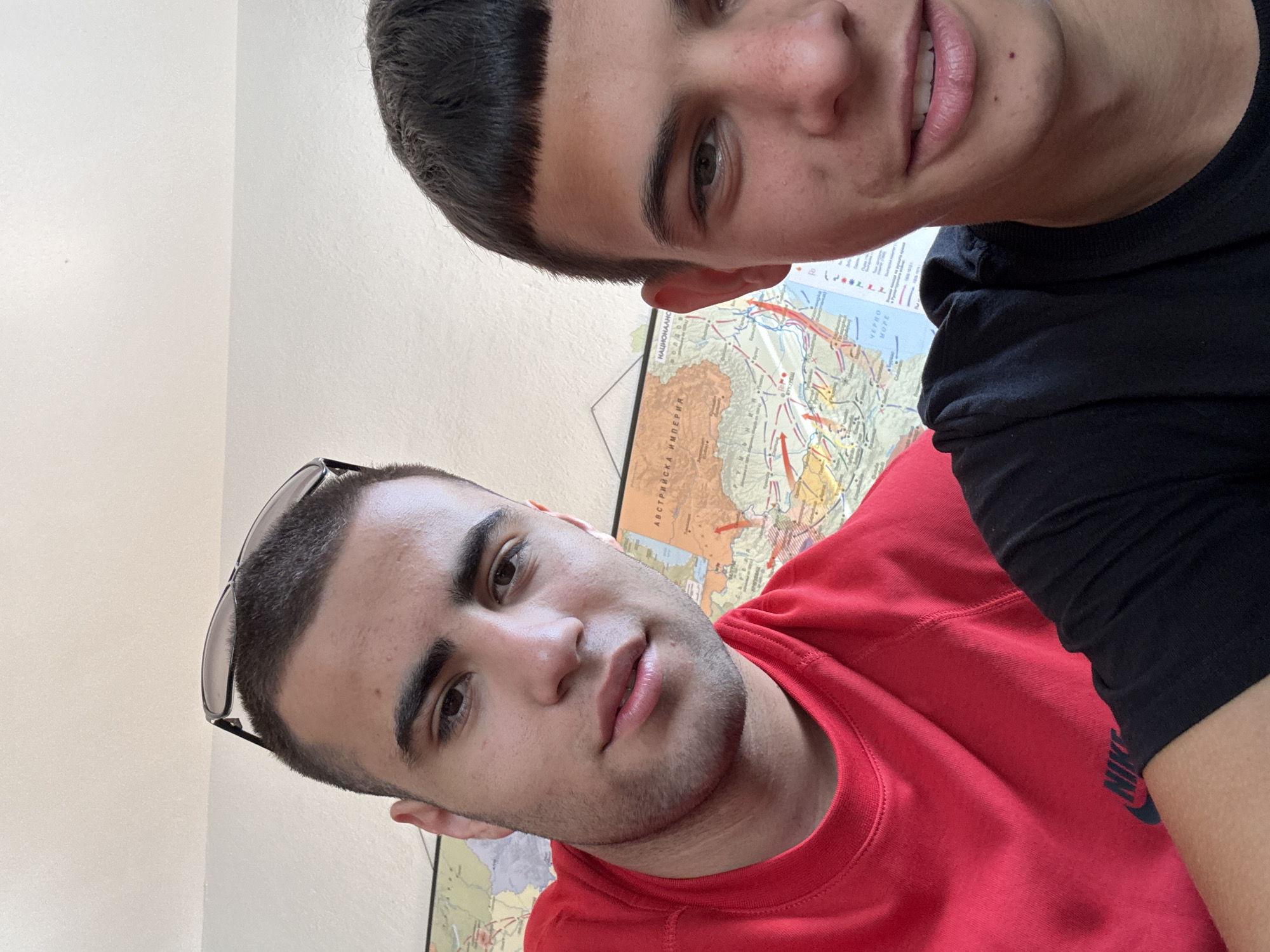The image shows two young men posing for a selfie in front of a wall with a map. The man on the left is wearing a red t-shirt and sunglasses perched on his head. The man on the right is wearing a black t-shirt. Both appear to be in good health and are looking at the camera. The map behind them shows a detailed political map of parts of Europe, with names in Cyrillic script, including "АВСТРИЙСКА ИМПЕРИЯ" (Austro-Hungarian Empire) and "НАЦИОНАЛНО" (National). The map also indicates some geographical features and possibly historical troop movements with red arrows. The overall environment appears to be indoors, possibly a classroom or an office, given the presence of the map. The lighting is bright, suggesting natural light from a window. The specific location within the city of Plovdiv, Bulgaria, cannot be determined from the visual information alone.