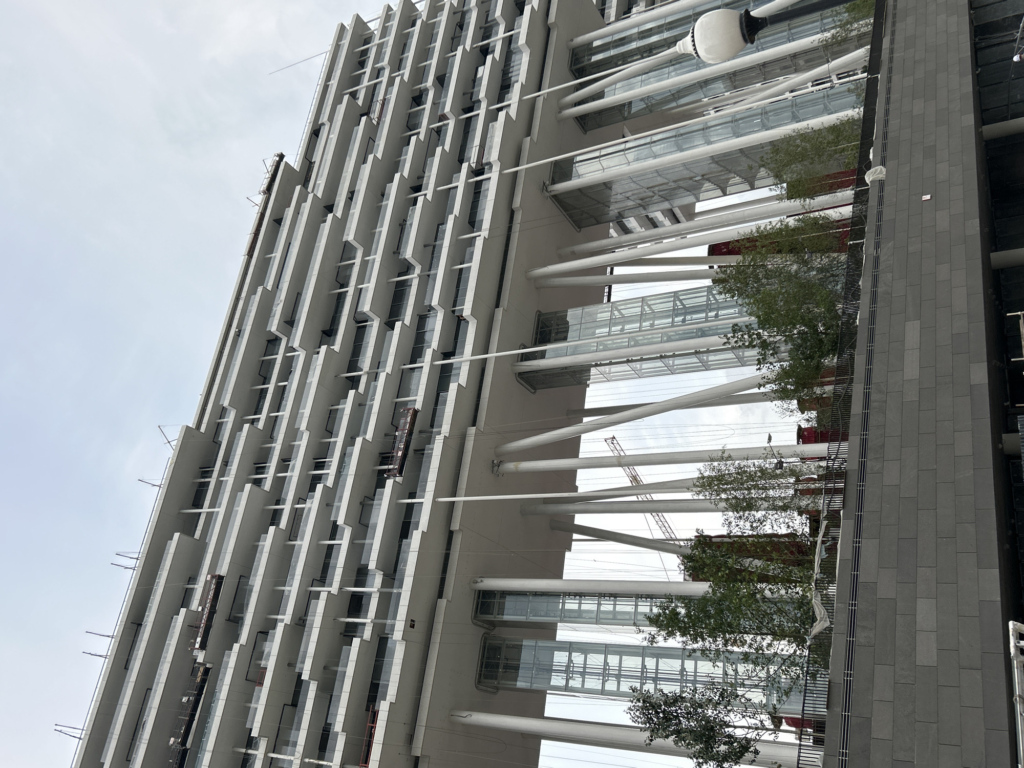 This image depicts a contemporary, multi-story building with a distinctive cantilevered design, characterized by numerous balconies and a series of white, angled support pillars beneath the main structure. The building appears to be under construction or recently completed, as evidenced by temporary scaffolding and materials visible on some of the upper levels.
The lower section of the building features tall, glass-enclosed elevator shafts and is partially obscured by trees and landscaping. A dark, textured stone facade forms the base of the structure in the foreground, with a metal railing running along the top of it. A vintage-style lamppost with a white globe is visible on the right side of the image. The sky above is overcast.
This building is located in Moscow, Russia, and is part of the "Dominion" business park, also known as "Moscow City" or "Moskva-Citi" architectural ensemble.