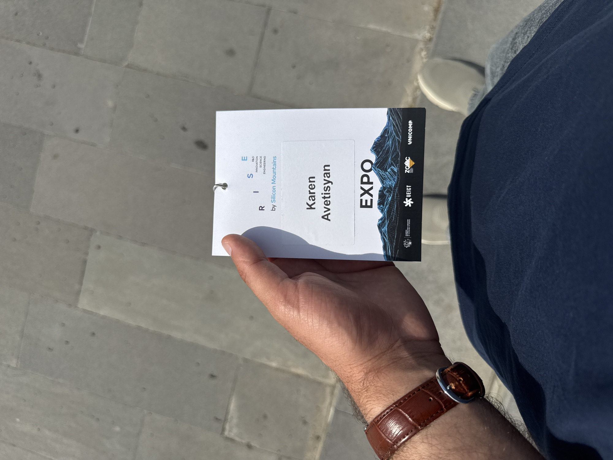 A first-person perspective photograph shows a person holding an event badge outdoors on a sunny day. The badge, predominantly white with black and blue design elements, features the name "Karen Avetisyan" prominently. Other visible text includes "EXPO" in large letters, and "RISE" with "R&D INNOVATION SCIENCE ENGINEERING by Silicon Mountains" below it. Several sponsor or partner logos are also visible on the badge: "UNICOMP", "ZCMC", and "*UEICT".
The person's right hand, wearing a brown leather wristwatch with a silver buckle, holds the badge. Part of their dark blue trousers and a light-colored sneaker are visible in the background. The ground beneath is a light gray concrete pavement, showing distinct seams and some darker marks, indicating an outdoor setting. Bright sunlight casts clear shadows of the hand and badge onto the ground, suggesting it is midday or early afternoon. The image captures a moment at what appears to be an expo, conference, or similar event.