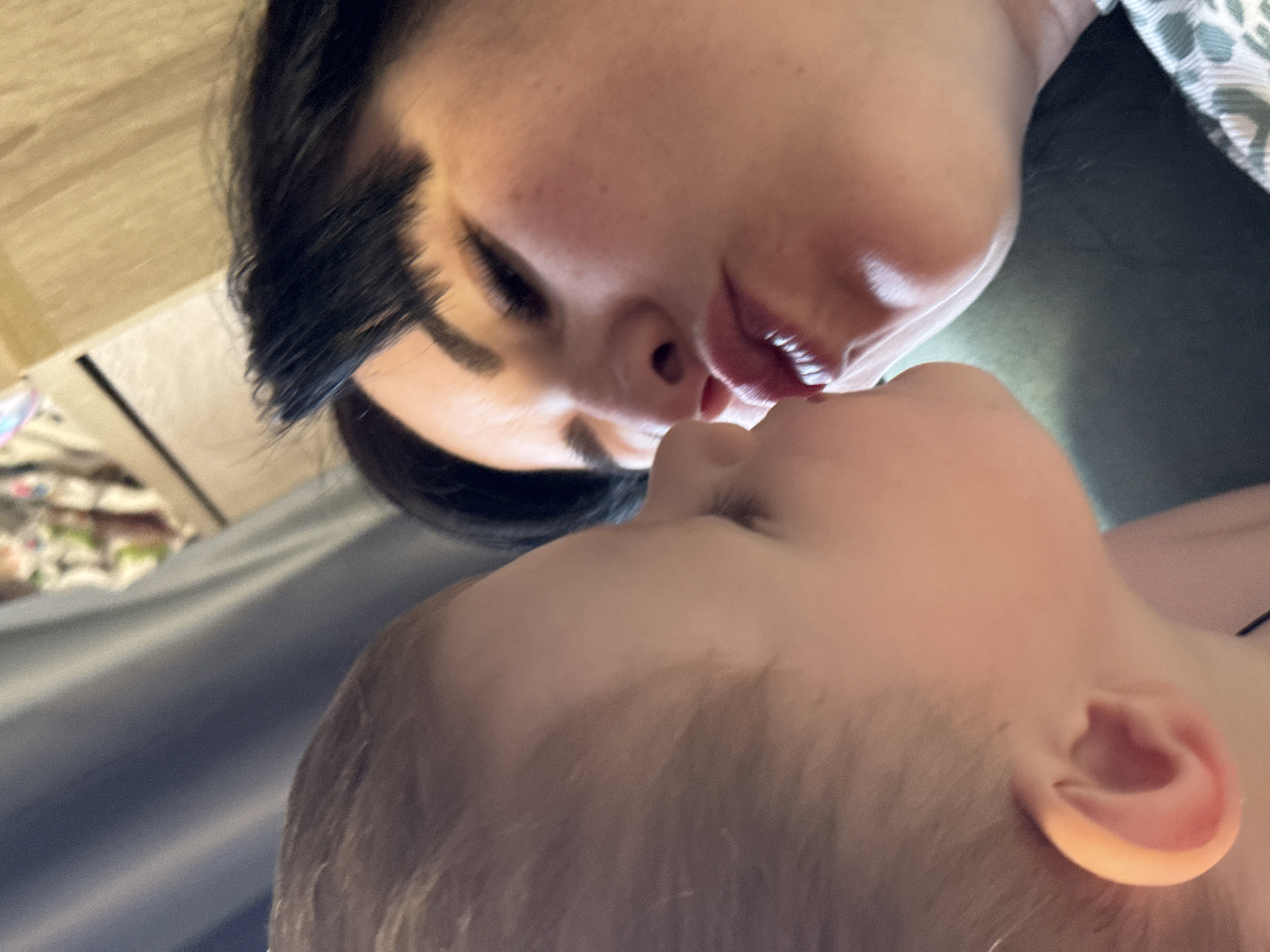 A close-up, upward-angle shot captures a woman and a child with their faces very near each other, possibly kissing or about to kiss. The woman has dark hair, fair skin, and her eyes are mostly closed. The child, positioned below the woman, has light-colored hair and a pink ear is visible. The background is blurry and includes indistinct elements suggesting furniture or bedding. There is no verifiable information to associate this image with the location in the city of Zav'yalovo, Russia.
