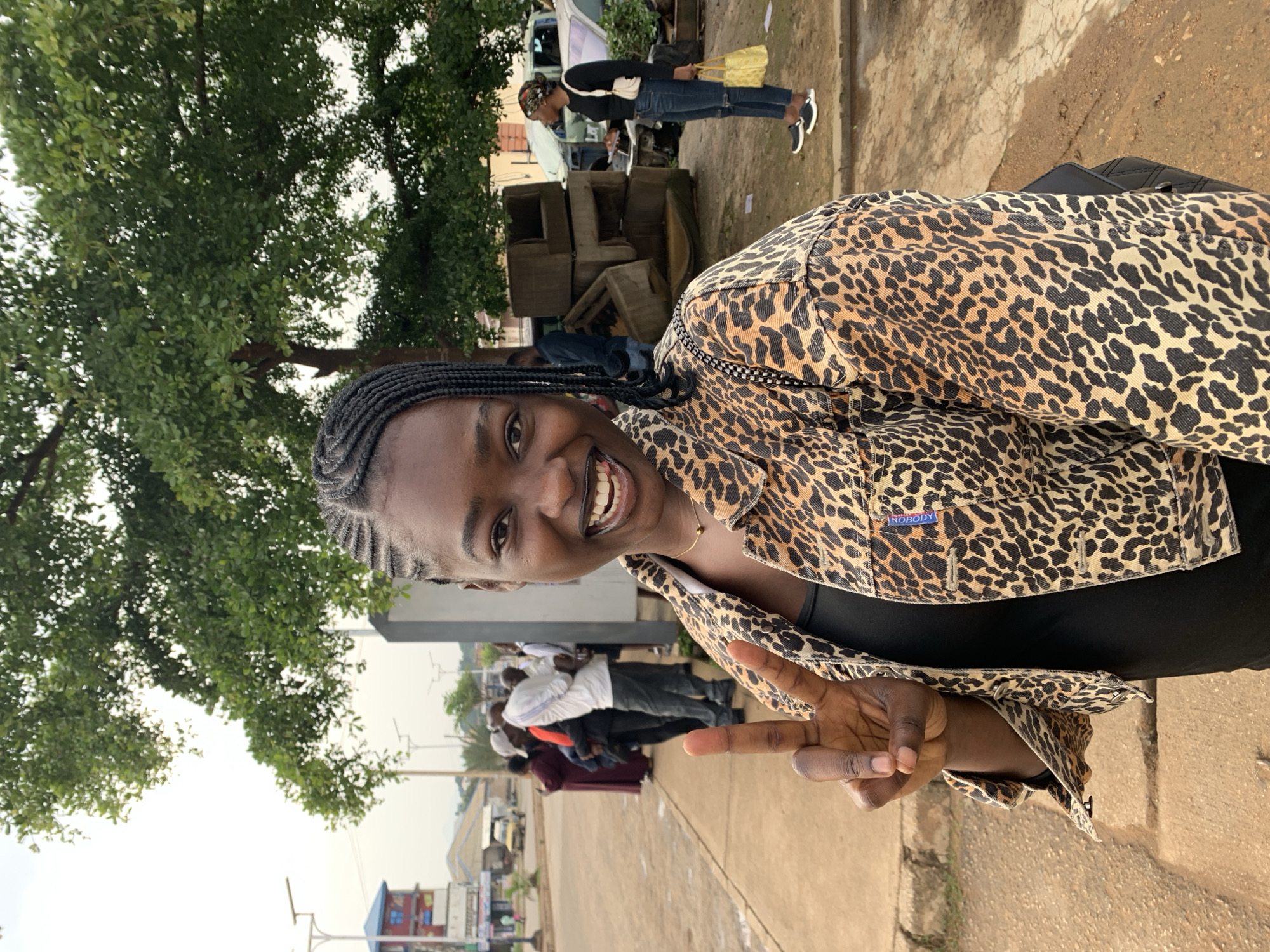 A medium shot photo shows a young Black woman in the foreground, smiling and holding up a peace sign with her left hand. She has her hair in braids and wears a leopard-print jacket over a black top. Behind her, on the right, a Black woman in a black shirt, blue jeans, and black slip-on shoes holds a yellow bag. Further in the background, several people are visible on a paved sidewalk, with trees and buildings suggesting an urban environment. The ground is a mix of dirt and pavement. The overall impression is that of a street scene, possibly in a market area. The photo has been geotagged to Rafin Par, Nigeria.