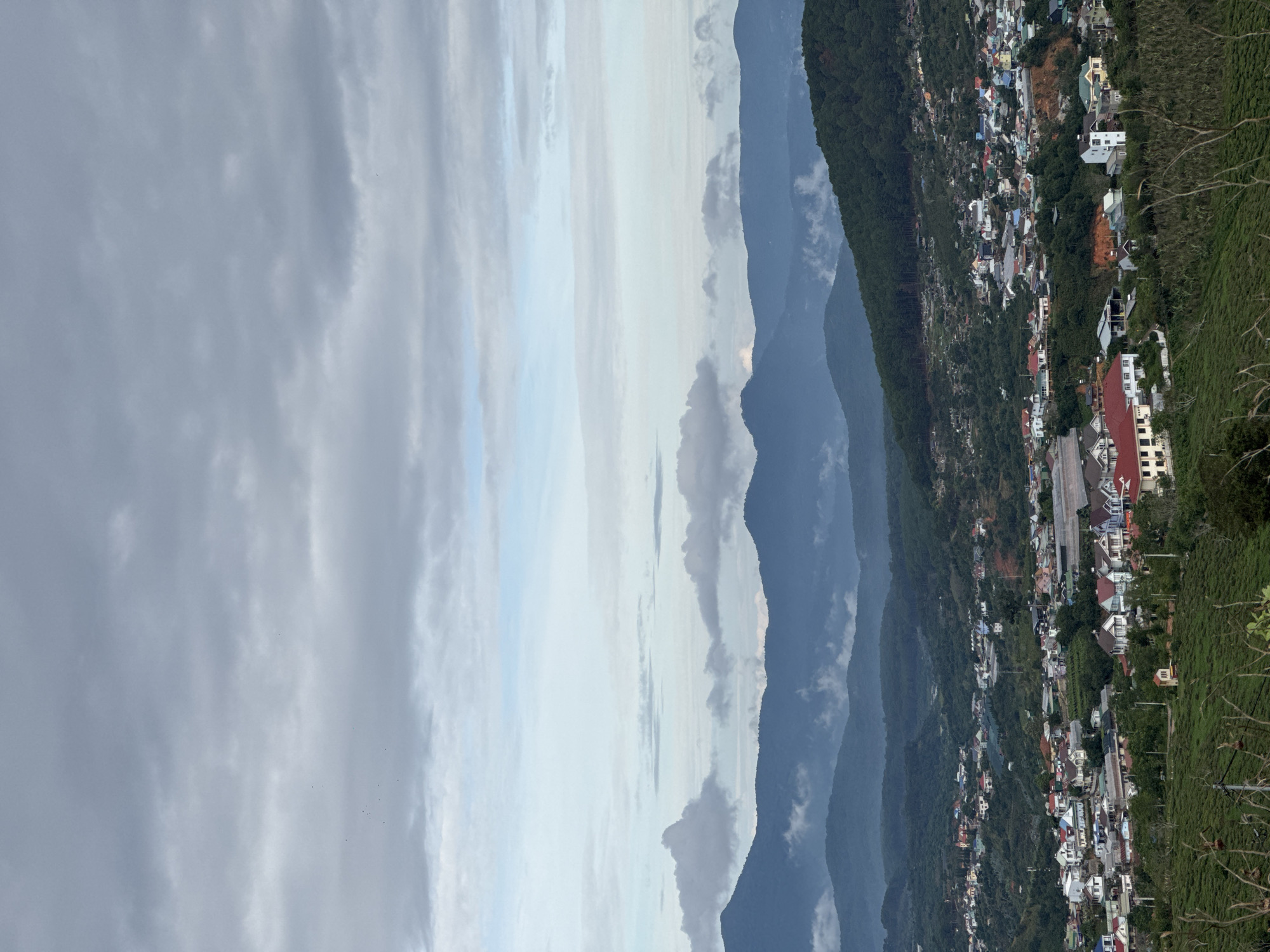 An elevated perspective showcases Trường Thọ, Vietnam. Multiple dark mountains line the center of the frame. A cloudy sky occupies the left side. The city is on the right, featuring clustered buildings interspersed with trees.