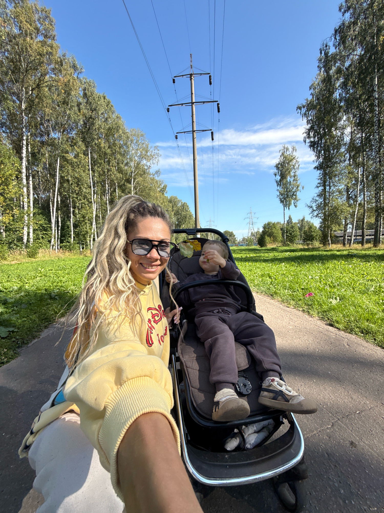 A medium-shot, eye-level selfie shows a woman with blonde, wavy hair pulled into a high ponytail, wearing large dark sunglasses and a yellow hoodie with red lettering. She is smiling and looking at the camera, with her left arm extended forward, holding the camera. Beside her, in a dark stroller, a young child in a dark gray outfit sits with their legs propped up on the stroller's footrest. The child's hands are near their face. The stroller is on a paved path that curves to the right, bordered by a grassy area on the left and right. In the background, a line of tall birch trees with green foliage runs along the left side of the path. Beyond the trees and across the grassy field, power lines and utility poles are visible against a clear blue sky with wispy white clouds. The setting appears to be a park or a pathway in a natural area, possibly in the city of Dumino, Russia, given the presence of birch trees and the clear blue sky.