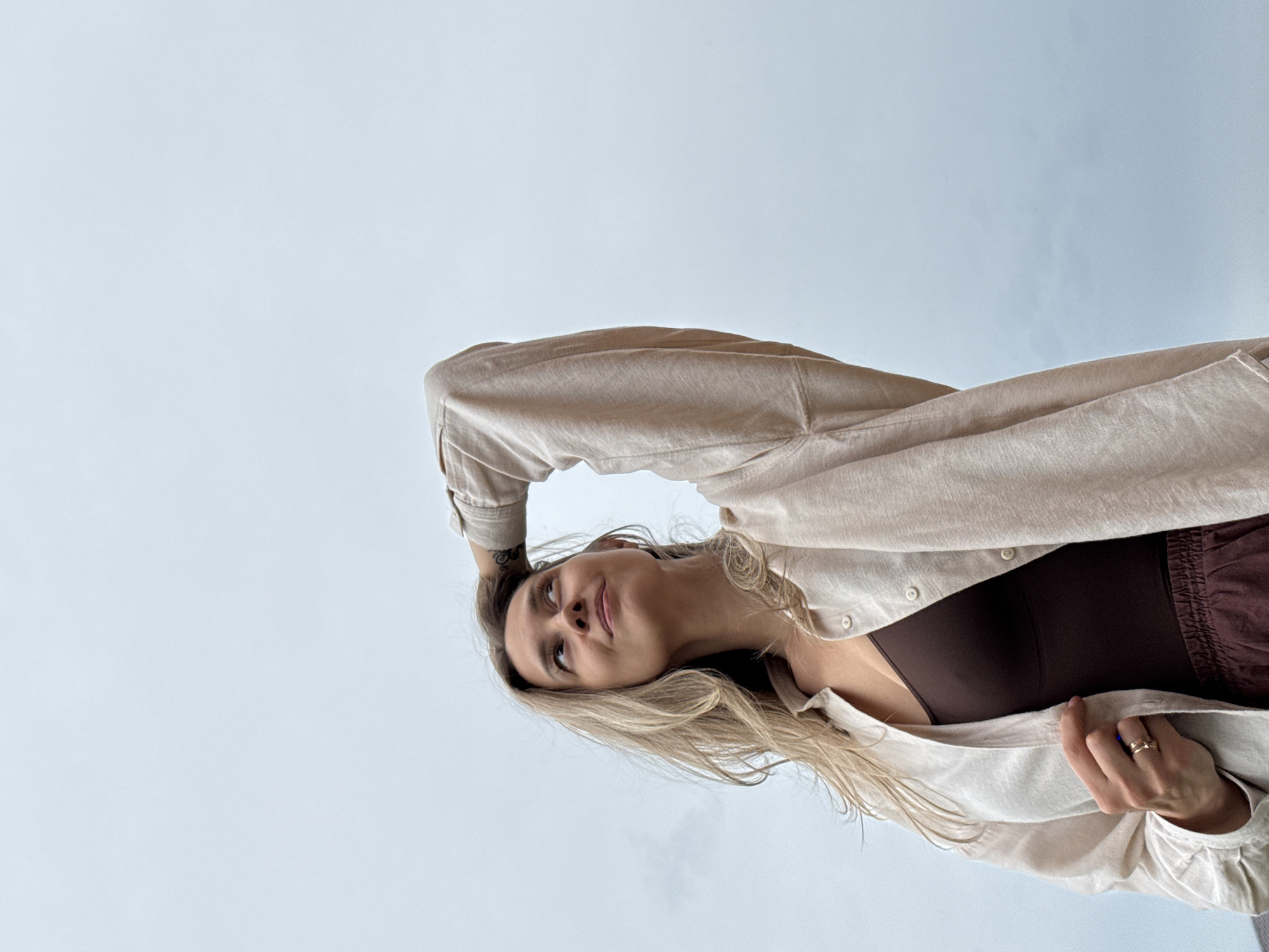 A low-angle medium shot captures a young woman with blonde hair, looking up and to the right, with her right arm raised and hand resting on her head. She is wearing a light-colored open button-up shirt over a dark brown tank top and reddish-brown pants. The background is a pale, overcast sky. The image appears to be taken outdoors with a muted color palette. No specific landmarks or distinguishing features are visible that would suggest a location within Chaikovsky, Russia.