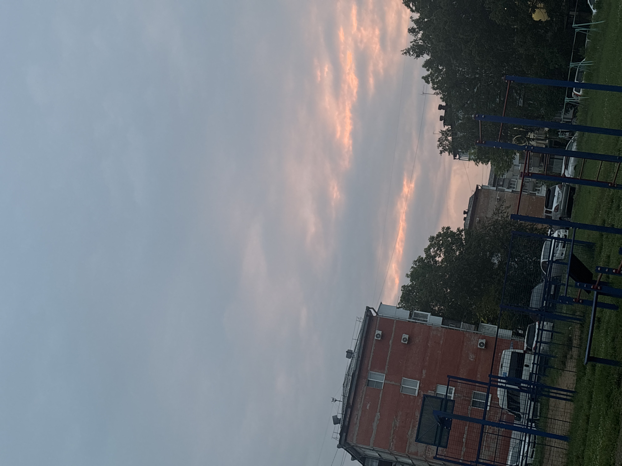 The image shows an outdoor scene in Birobidzhan, Russia, at dusk or dawn. The sky is filled with pale blue and pink clouds. In the foreground, there is a playground with blue metal bars and green grass. Behind the playground, there are several parked cars, including a white van. Further in the background, there are apartment buildings and trees. The apartment buildings appear to be made of brick and concrete, and some have air conditioning units visible on their facades. The overall impression is of a residential urban area.
