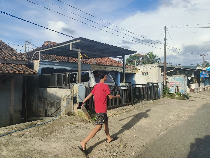 The image shows a young man walking away from the camera, in a red shirt and blue jeans, holding a black cell phone in his right hand. He is walking towards a building with a tin roof, with the street behind him leading to a small town. The location is Cikalong, Indonesia, with coordinates -6.758 latitude and 107.447 longitude. The image was taken on a bright, sunny day, with blue skies and white clouds. The ground is dusty and uneven. There are power lines overhead.