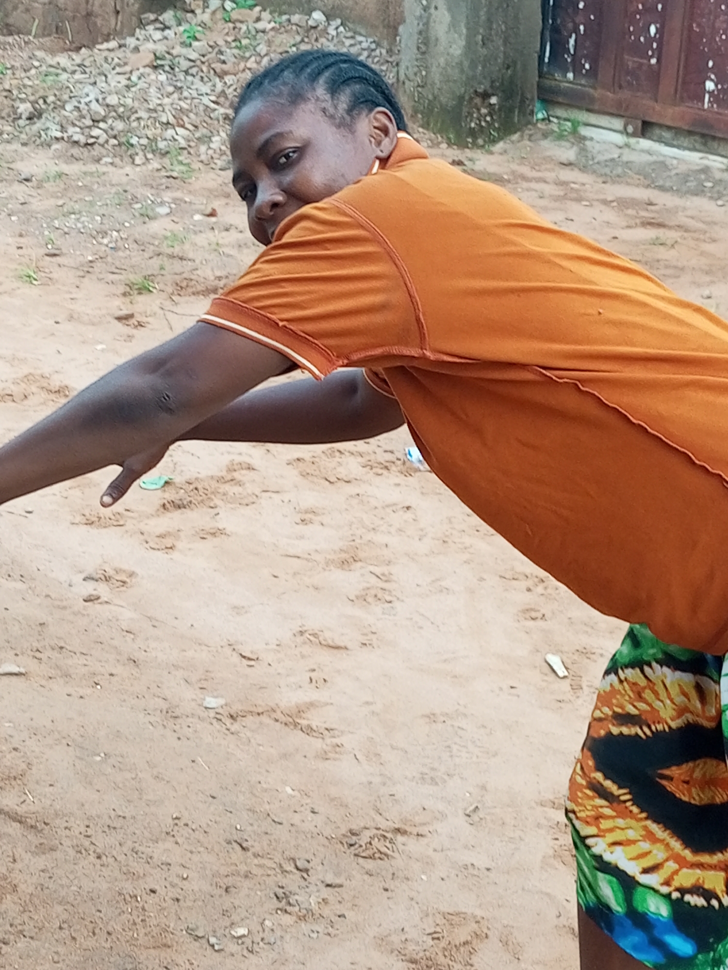 A medium shot captures a young Black woman in a crouched position, outdoors in what appears to be a sandy area with gravel and some sparse vegetation. The woman is wearing an orange t-shirt and a patterned skirt with green, yellow, and black designs. Her face is turned towards the camera, and she is looking directly at the viewer with a neutral expression. Her arms are extended forward, with her right hand resting on the sandy ground. The background is out of focus, showing a wall and what might be a fence or gate. The lighting suggests it is daytime, with a slightly overcast sky.