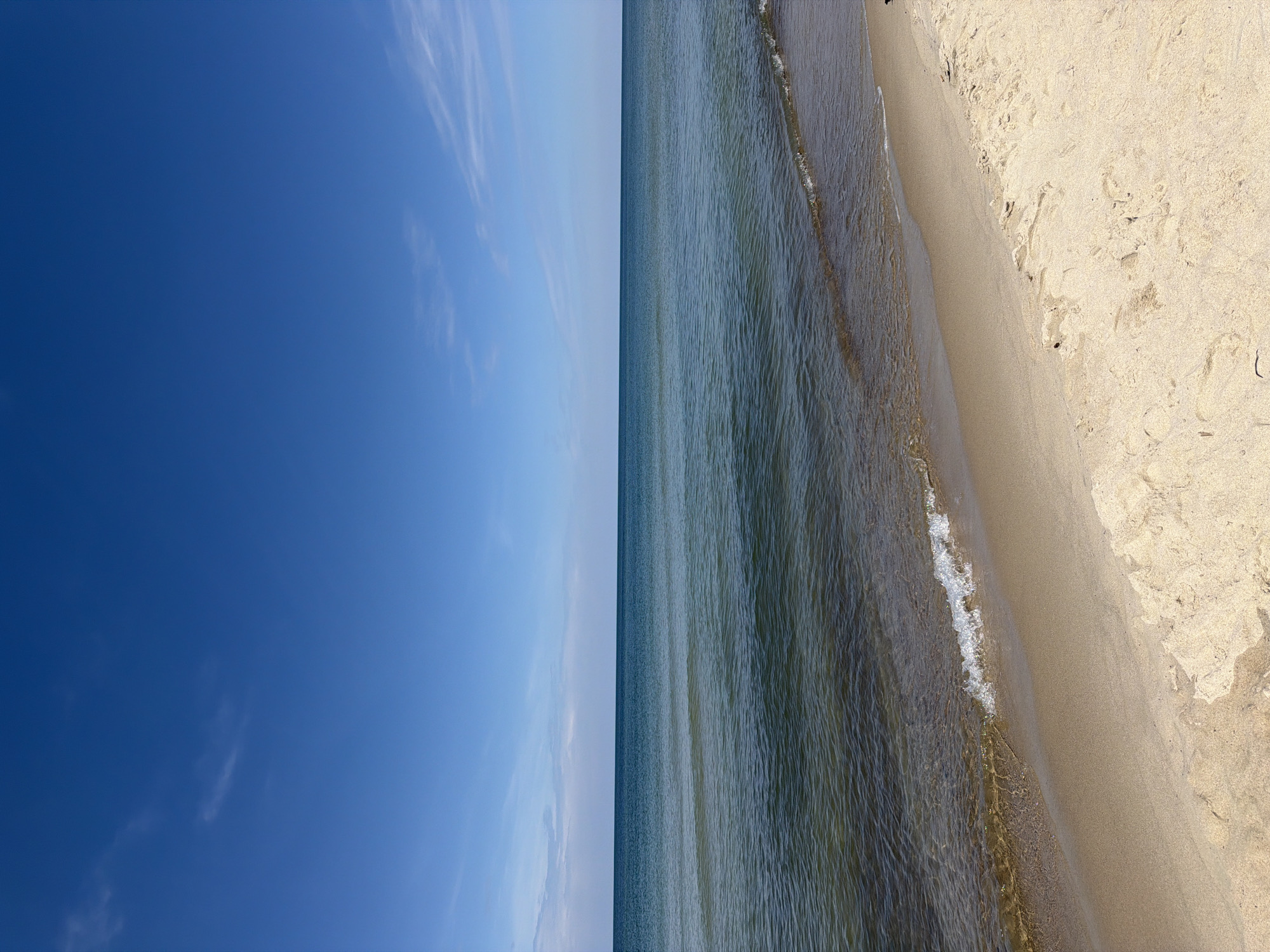 The image shows a serene beach scene on a clear day. The foreground is dominated by light-colored sand, with some subtle footprints visible. A gentle wave, with white foam, is washing ashore, creating a clear line between the wet and dry sand. Beyond the sand, the calm, shallow water transitions to a deeper, bluish-green hue. The horizon line is sharp and distinct, separating the sea from a clear blue sky. A few wispy clouds are scattered across the upper portion of the sky. The scene depicts the natural beauty of a coastal environment, likely the Baltic Sea coast near Morskoye, Russia, given its tranquil and expansive appearance. There are no people or man-made objects clearly visible in the frame, contributing to the peaceful atmosphere.