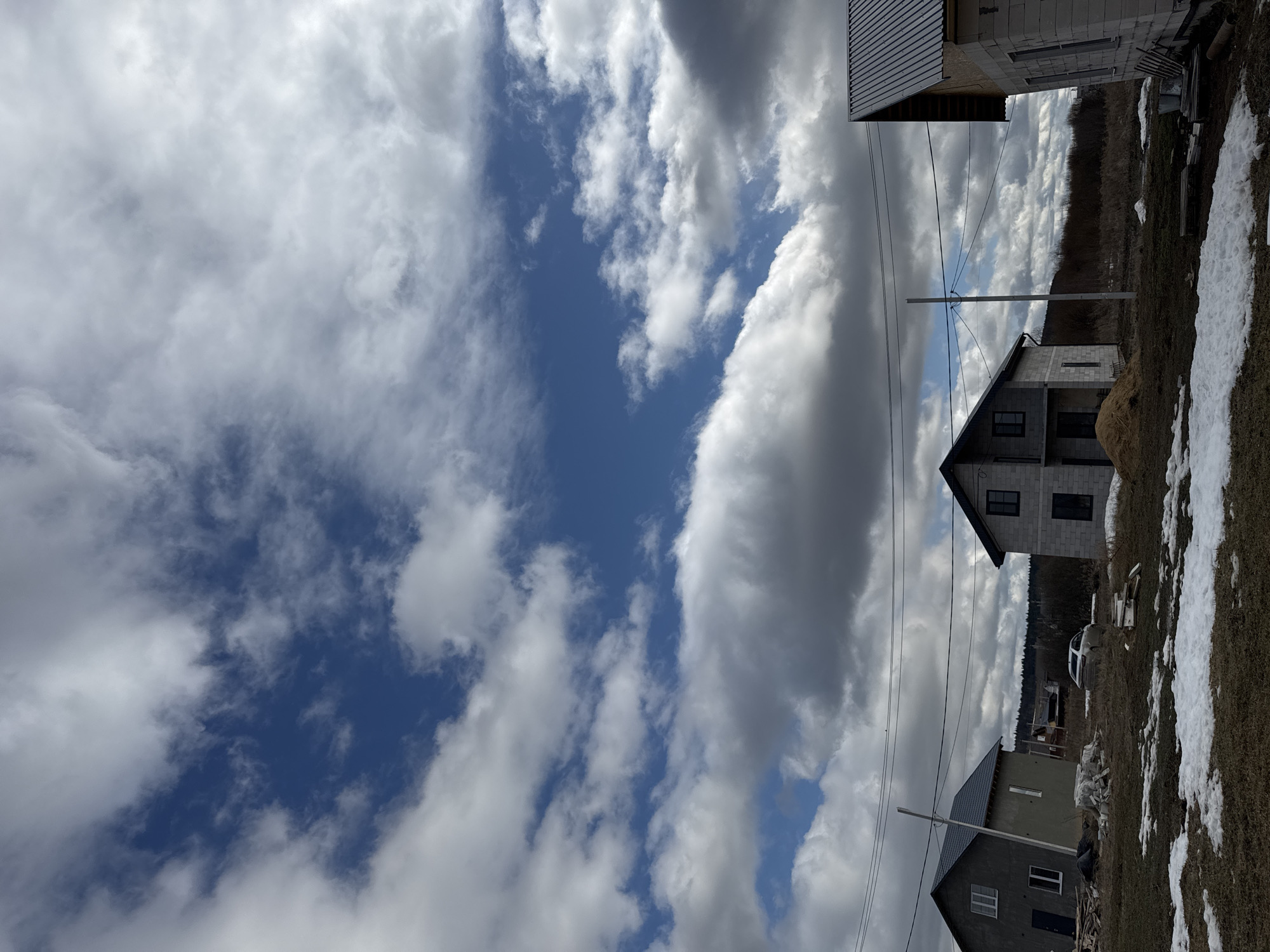 The image shows a partially cloudy sky with a view of construction in Gornyi, Russia. Two unfinished, grey, two-story houses with dark-colored roofs are visible. A utility pole with power lines runs across the scene. Patches of melting snow and dry grass cover the ground. In the background, there's a line of trees. A car is parked near one of the houses, and there are construction materials scattered around the area.