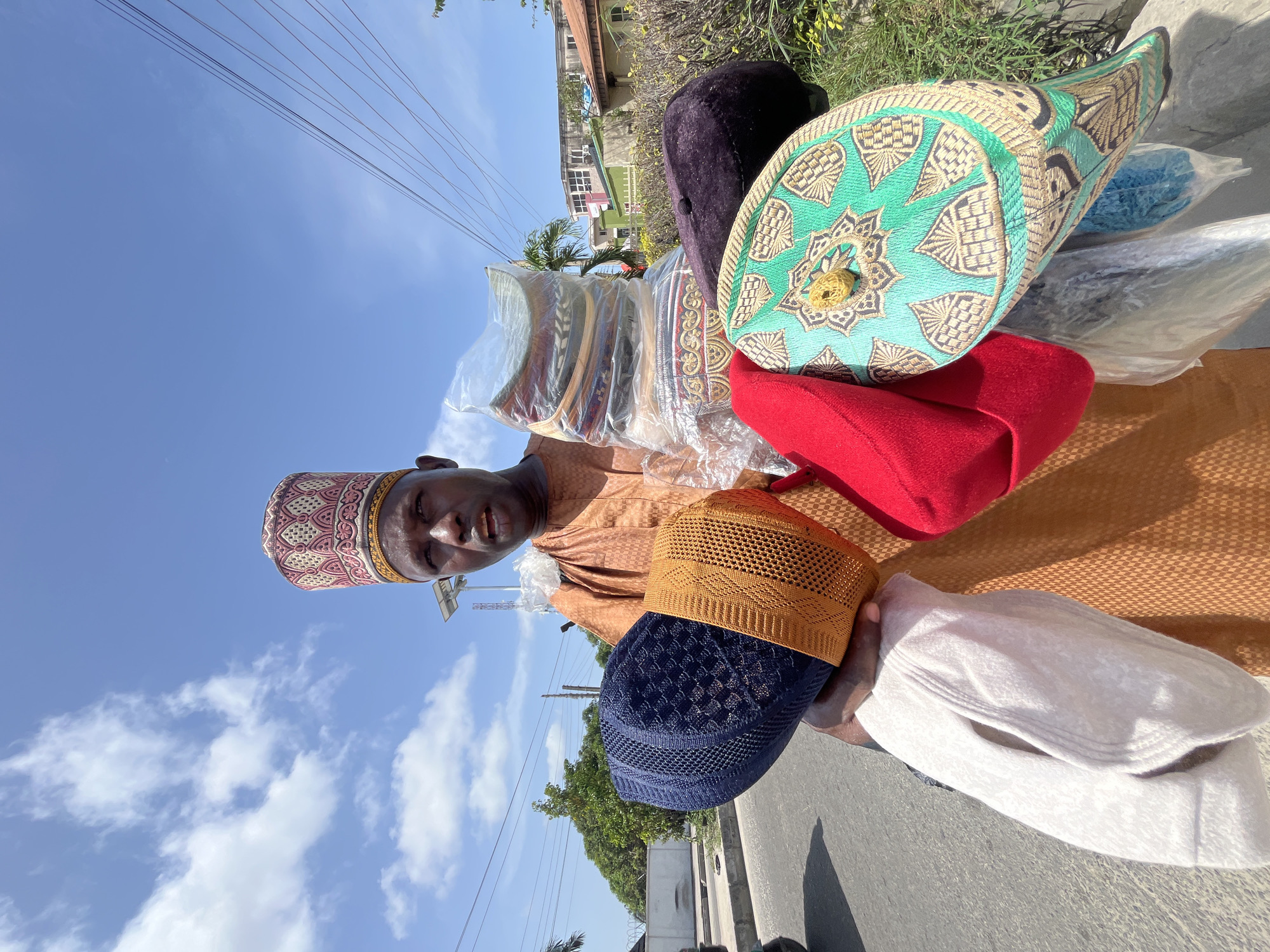 A man stands on the side of a road in Ado, Nigeria, holding a collection of various caps, most likely for sale. He wears a brown patterned robe and a patterned cap. He holds caps of multiple colors and styles, including red, blue, white, green, purple, and yellow. A stack of caps is wrapped in plastic. The background shows a bright blue sky with clouds, power lines, and parts of buildings and vegetation. The setting appears to be an urban roadside.