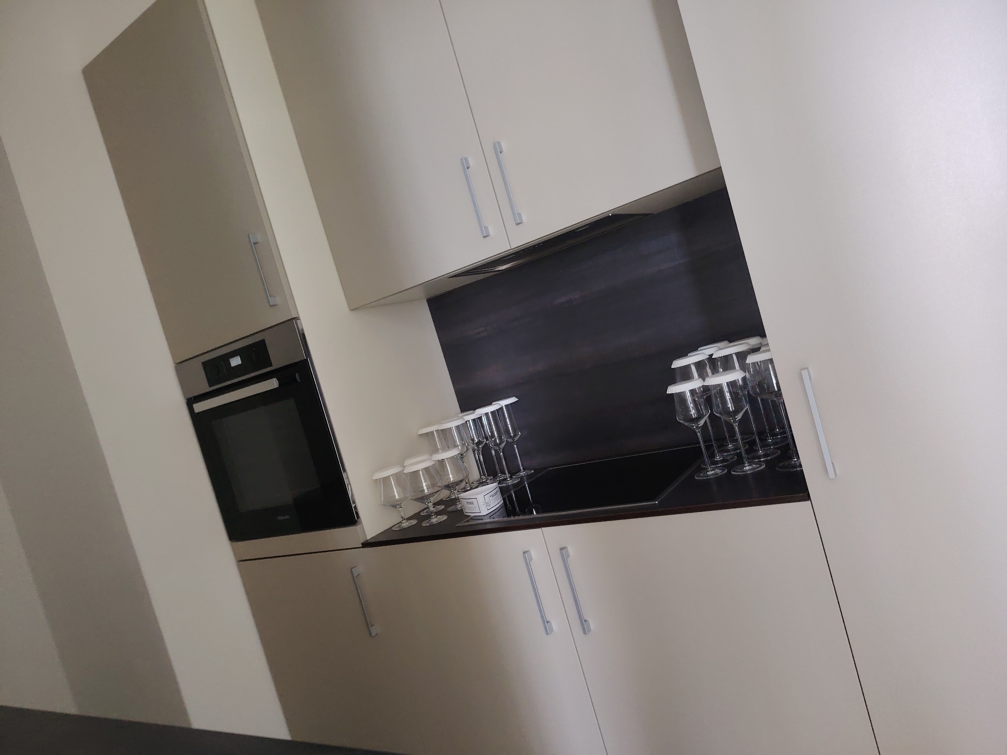 The image displays a modern kitchen interior with light-colored cabinets and a dark backsplash. The kitchen features an oven on the left, integrated into a cabinet. On the countertop, several wine glasses with white stoppers are arranged in rows, next to a black induction cooktop. Two cabinet doors with silver handles are visible on the lower part of the image. The overall scene is clean and organized, suggesting a domestic or commercial setting. The image is captured from a slightly tilted perspective.