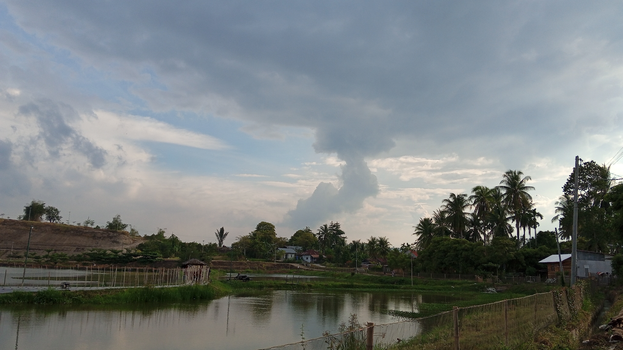 A wide shot shows a rural landscape in Geulangganggampong, Indonesia, under a cloudy sky. In the foreground, a calm body of water reflects the overcast sky. A low wooden fence with thin poles runs along the edge of the water, partially enclosing an area with green grass and some scattered vegetation. Beyond the water, a line of trees, including several palm trees, forms a natural barrier, with some houses nestled among them. To the left, a sloped dirt embankment is visible, dotted with sparse vegetation and a few trees. In the mid-ground, a small, rustic wooden hut with a thatched roof stands near the water's edge, and a fence extends from it. Further back, more houses and a cluster of dense green foliage are visible. The sky is dominated by heavy, dark clouds with patches of lighter sky peeking through.