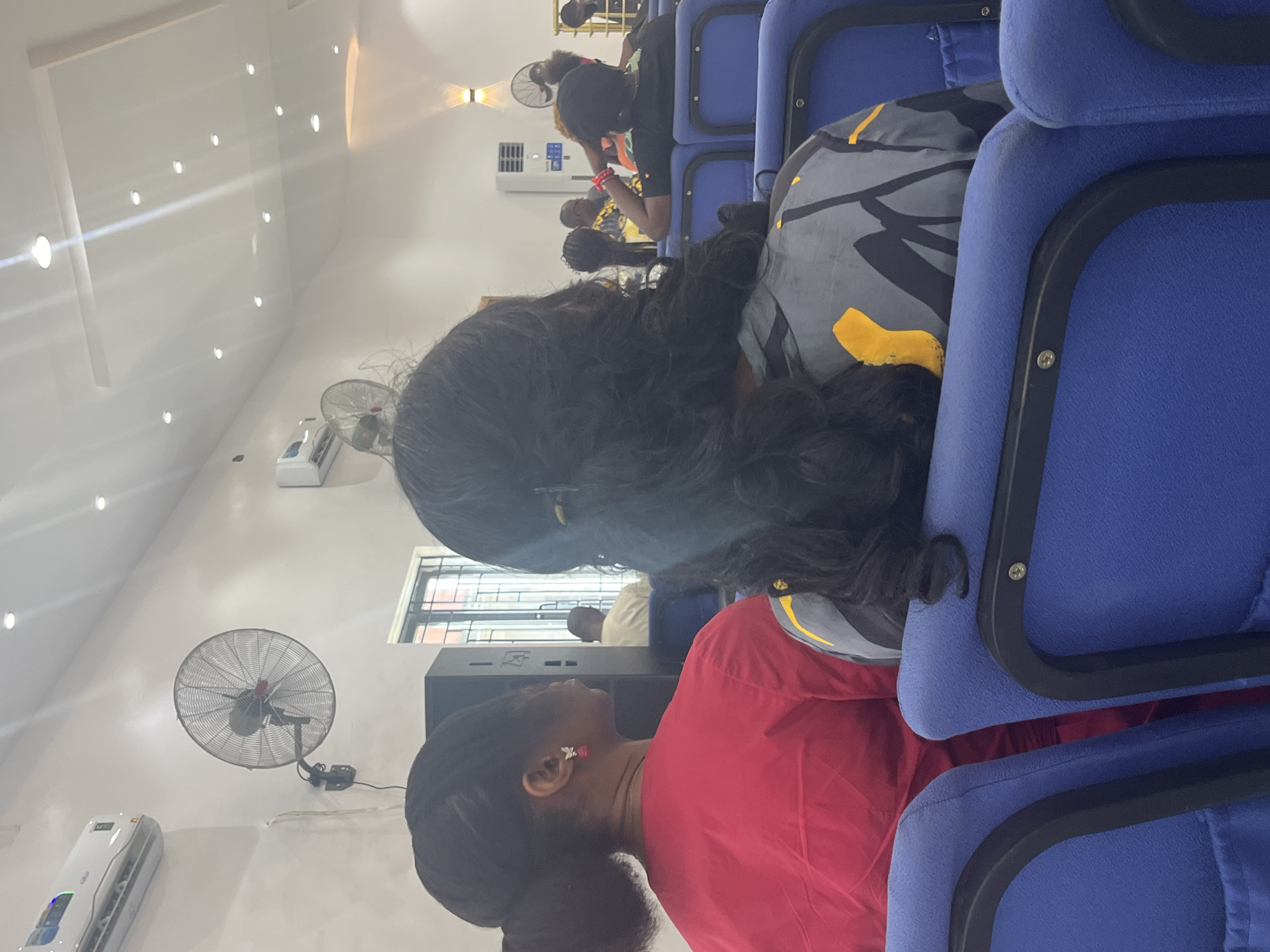 The image depicts a scene from within what appears to be a church in Ado, Nigeria. Rows of blue upholstered chairs are visible, occupied by several individuals. The foreground shows the back of a woman with dark hair and a red top. To her left, there is a speaker. In the background, there are other people, various fans and lights. The environment has a modern, clean appearance.