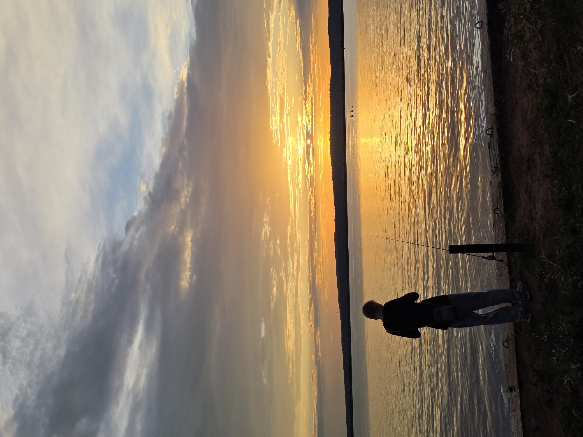 A lone individual stands on the shore of a wide body of water, facing away from the camera and towards a vibrant sunset. The sky is a spectacle of oranges, yellows, and soft purples, with dramatic, textured clouds. The water reflects the warm hues of the sunset, creating a shimmering, golden surface. In the distance, a faint dark line of hills or land breaks the horizon. The foreground shows the person, dressed in a dark top and light-colored pants, holding a fishing rod. A fishing rod is also propped up nearby. The ground beneath the person is a mix of dark earth and sparse vegetation. The overall atmosphere is one of peaceful solitude during a beautiful natural event, possibly a sunset over the Kama River near the city of Chaikovsky, Russia, known for its expansive water reservoirs.