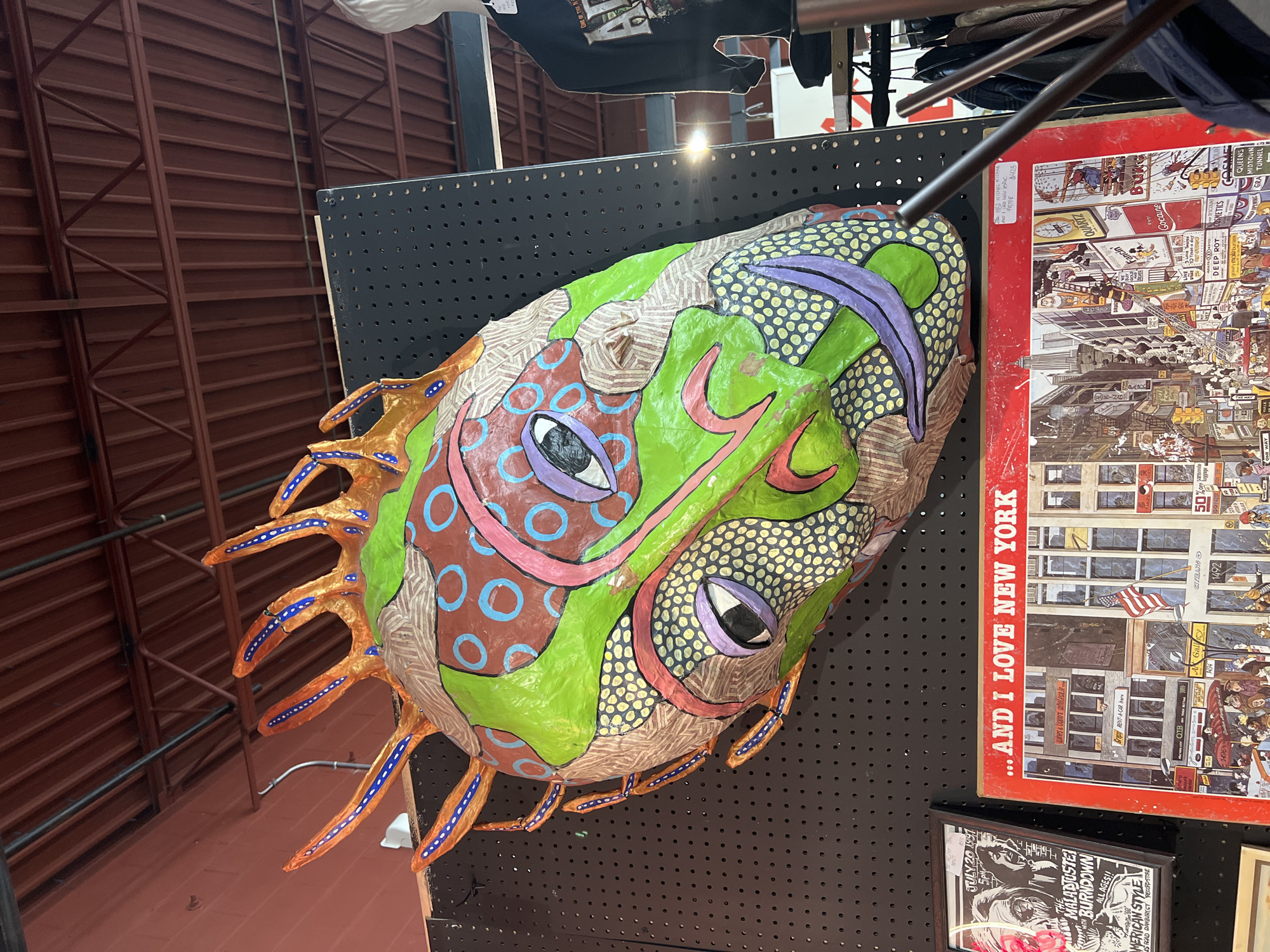 The image shows a colorful, paper mache mask of a face with exaggerated features hanging on a black, perforated display panel. The mask has protruding, tentacle-like appendages. The background reveals a section of a red roof structure and clothing hanging above the display panel. To the right of the mask is a poster with a red border, densely packed with cartoonish depictions of buildings and signs, including "AND I LOVE NEW YORK" printed prominently. Below the New York poster is a black and white poster, which contains text indicating "July 26 1991" as well as other miscellaneous poster ads. The location, City of Newburgh, United States of America is not identifiable from the image.