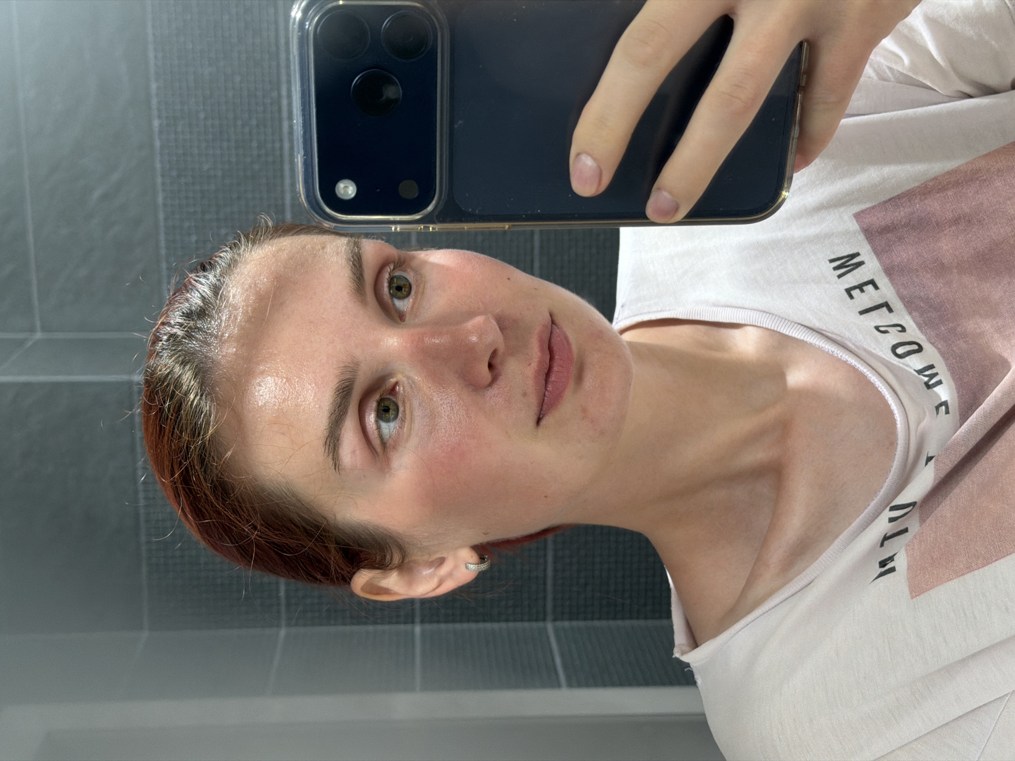 A woman takes a selfie in a bathroom. She holds an iPhone in front of her face. She wears a white T-shirt with a pink geometric design and lettering. The background shows gray tiled walls. The location in the city of Chaikovsky, Russia is unknown.