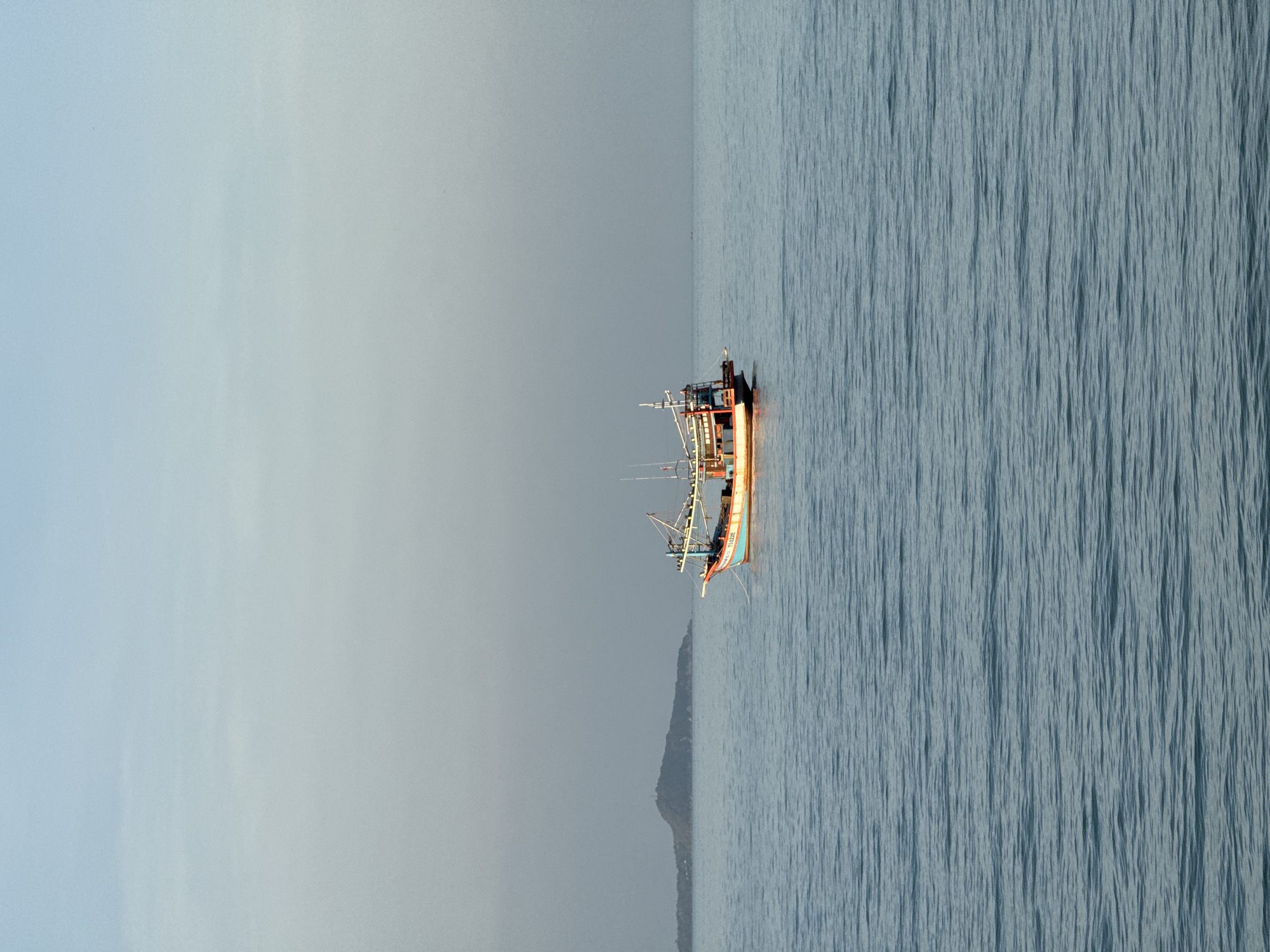 A fishing boat is anchored in the calm, blue sea off the coast of Ko Samui District, Thailand. The boat, which is painted in vibrant colors of red, orange, and blue, has a traditional design with a tall mast and rigging. In the background, a lush green island with a gentle slope rises from the water, shrouded in a light haze. The sky above is a pale blue, transitioning into a soft grey near the horizon, suggesting either early morning or late afternoon light. The water is gently rippled, reflecting the subdued colors of the sky and the boat.