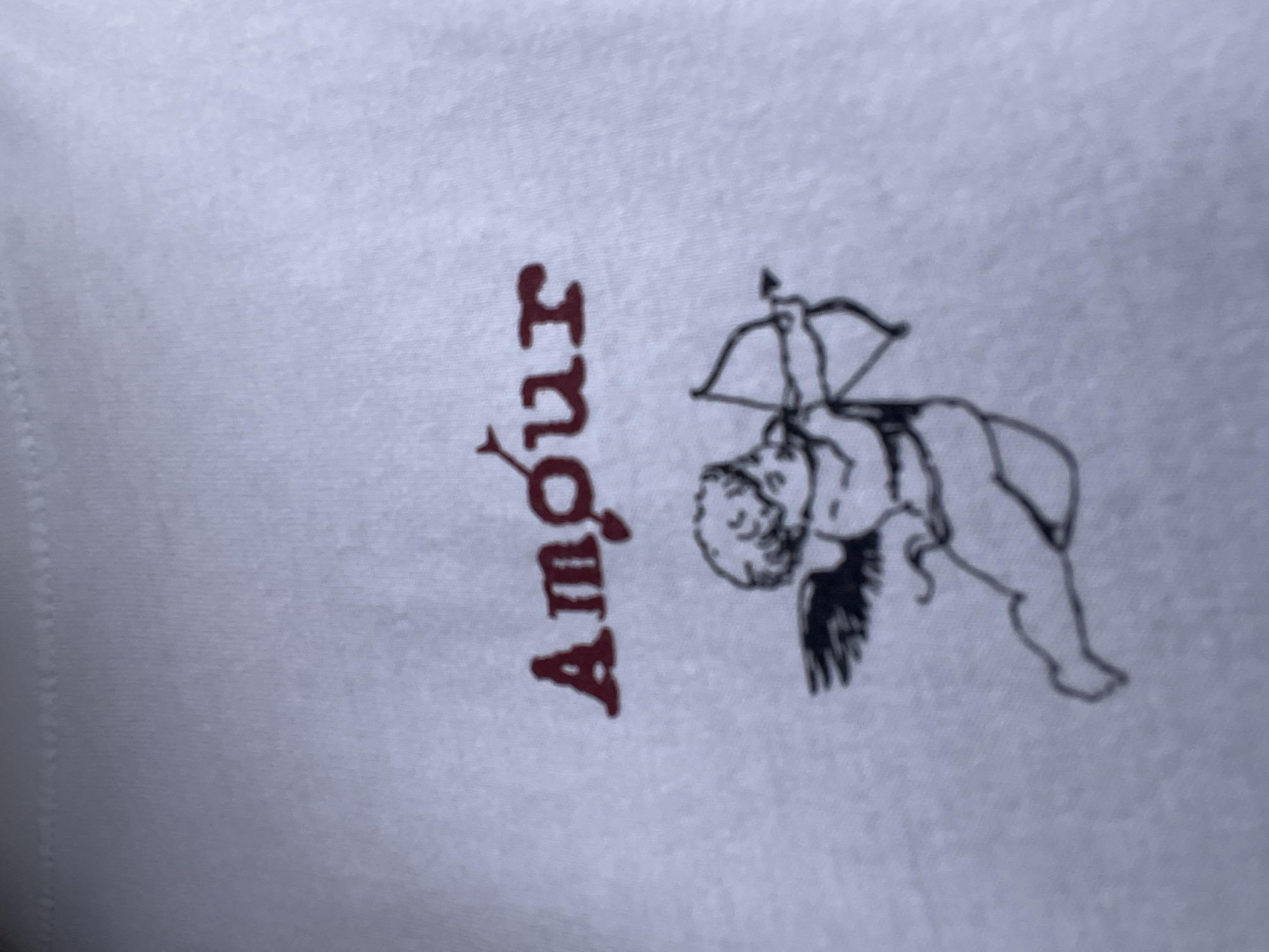 This media file contains a close-up of a white fabric, likely clothing or a textile, with a decorative graphic printed on it. The graphic features a drawing of a cherubic figure, similar to Cupid, with wings and a bow and arrow. The cherub is depicted in a dynamic pose, leaning forward with its bow drawn. To the left of the cherub, the word "Amour" is printed in a dark red, stylized font, with the letters oriented vertically and upside down. The image is slightly blurry, and the fabric appears to have a subtle texture. There are no discernible cues about the time of day, weather, or specific emotions conveyed beyond the theme of love suggested by the word "Amour" and the imagery of Cupid. The location context of Chaikovsky, Russia, does not provide any visual cues within the image itself.